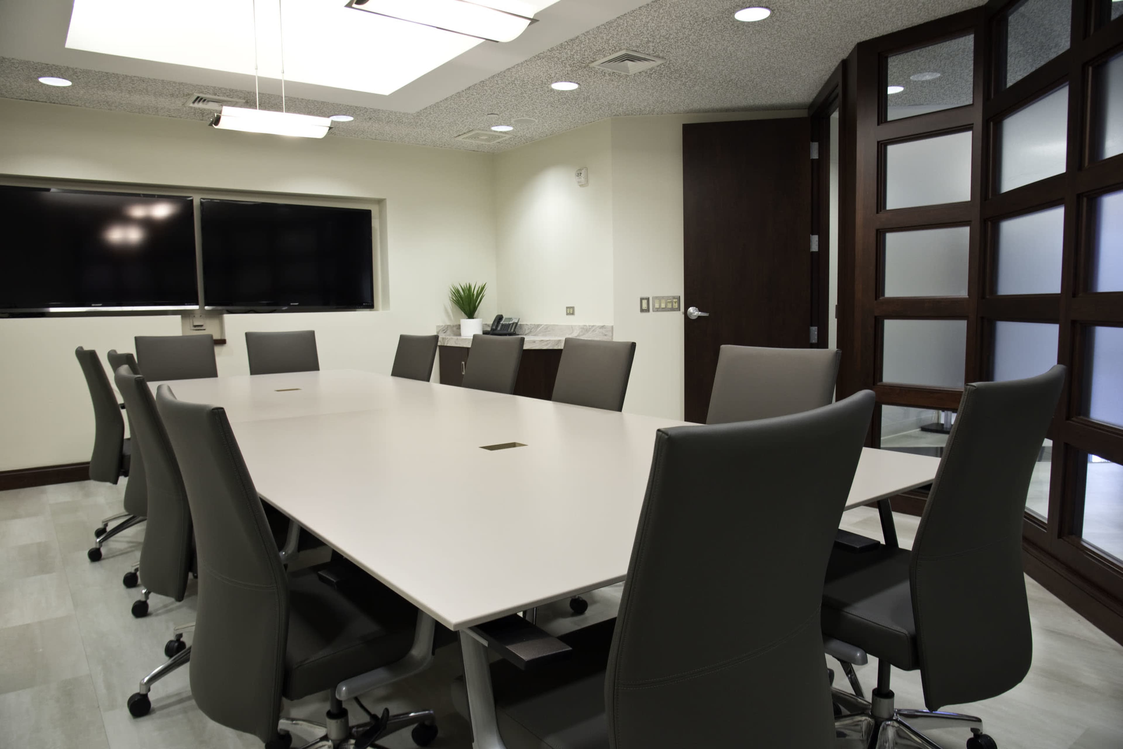A modern conference room features a large white table surrounded by ergonomic chairs, with a wall-mounted screen and a glass-paneled door.