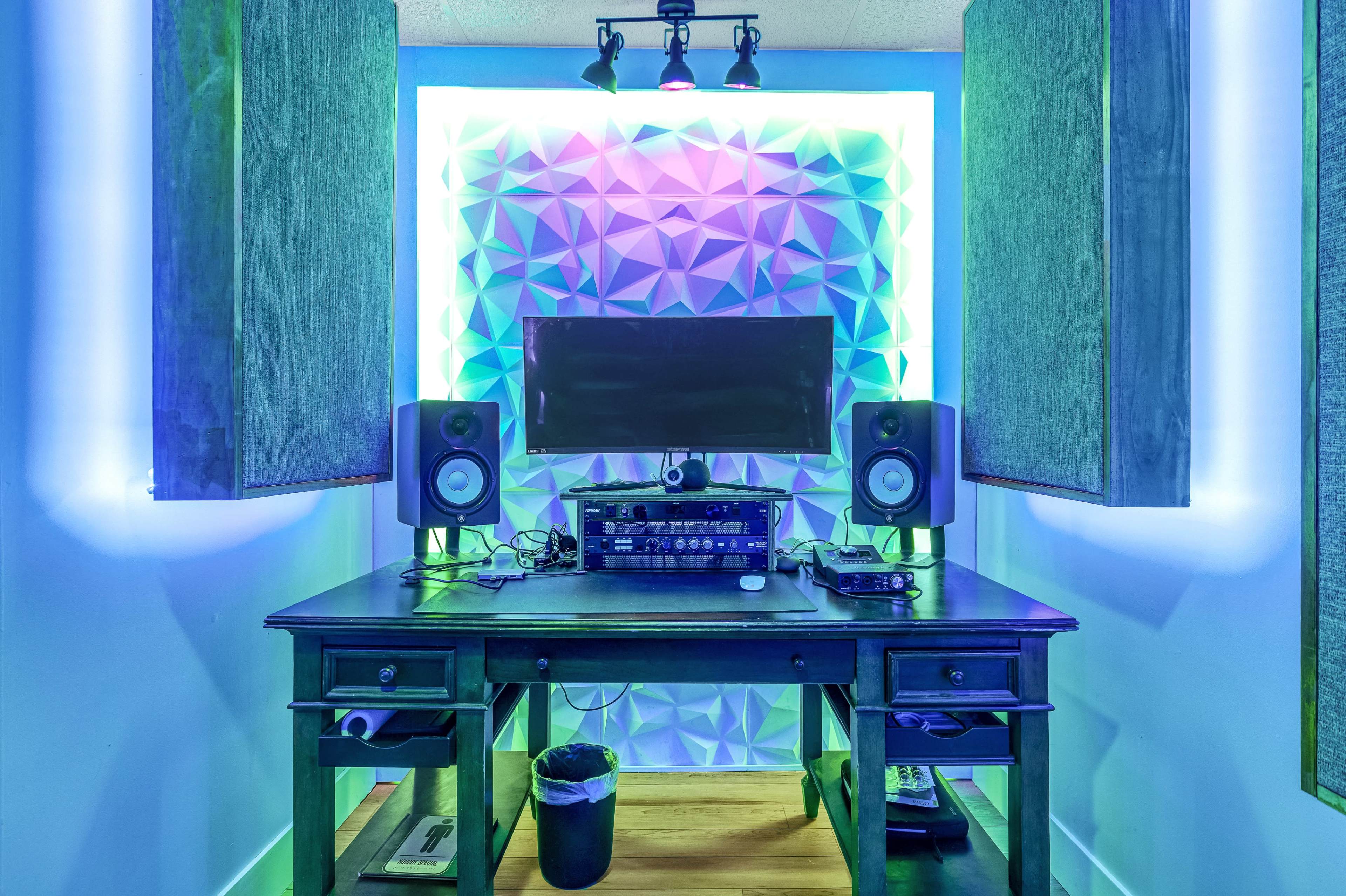 A home recording studio featuring a desk with audio equipment, two speakers, and a monitor, illuminated by colored LED lights against a textured wall.