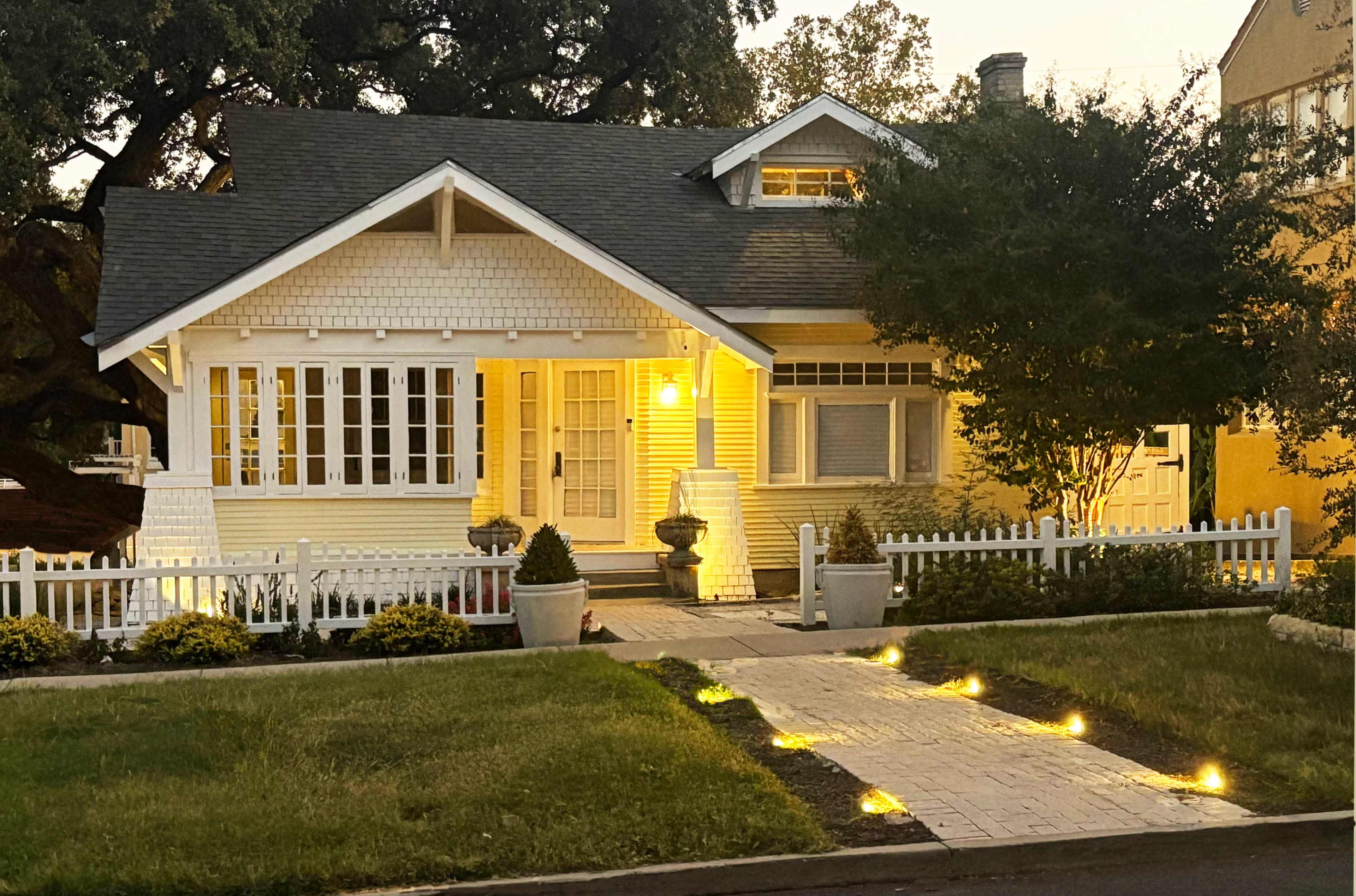 A well-lit, charming bungalow with a white picket fence and garden path lined with lights.