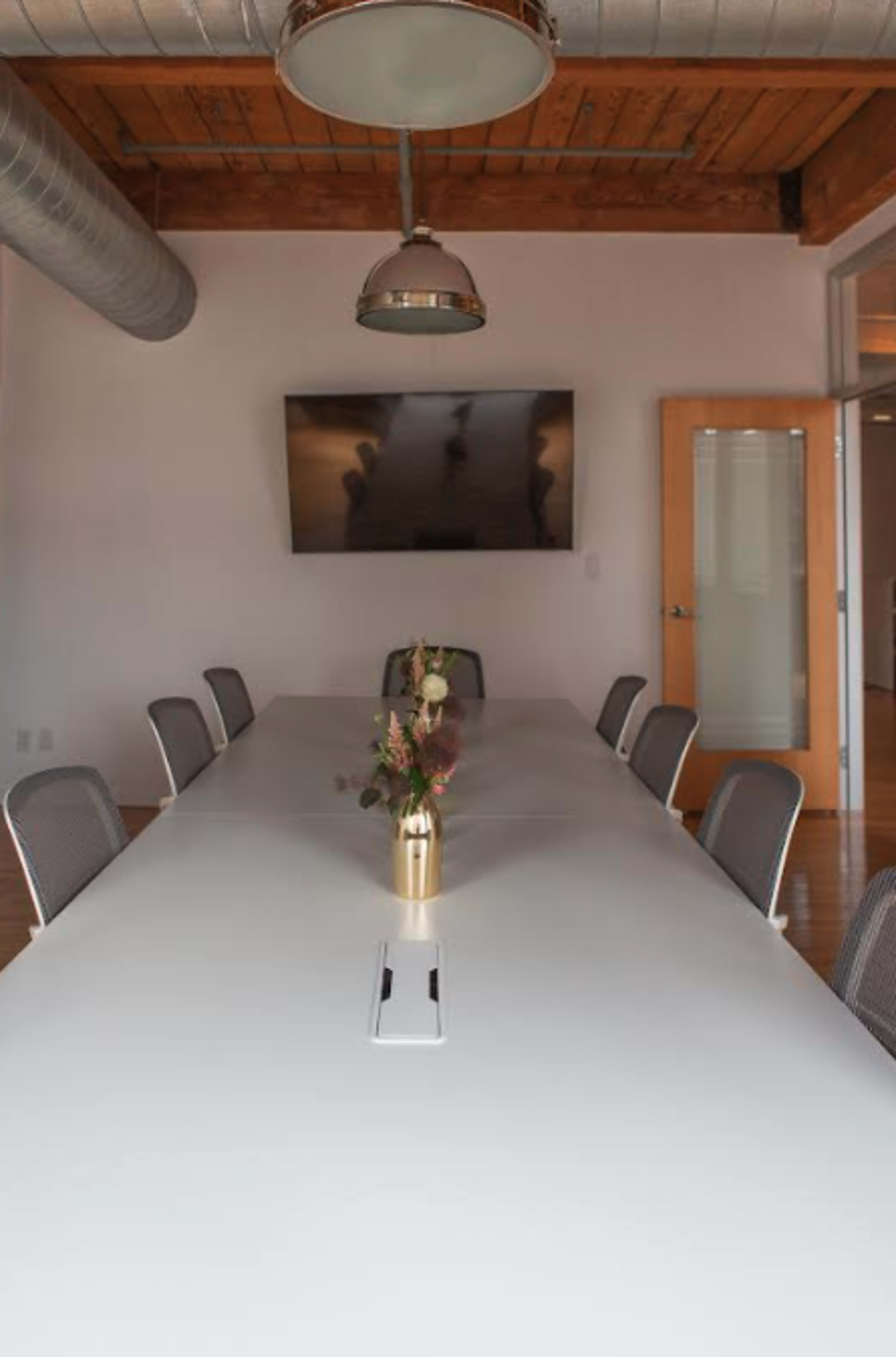 A long, white conference table is set in a meeting room with metal chairs and a television mounted on the wall, featuring a small vase of flowers at its center.