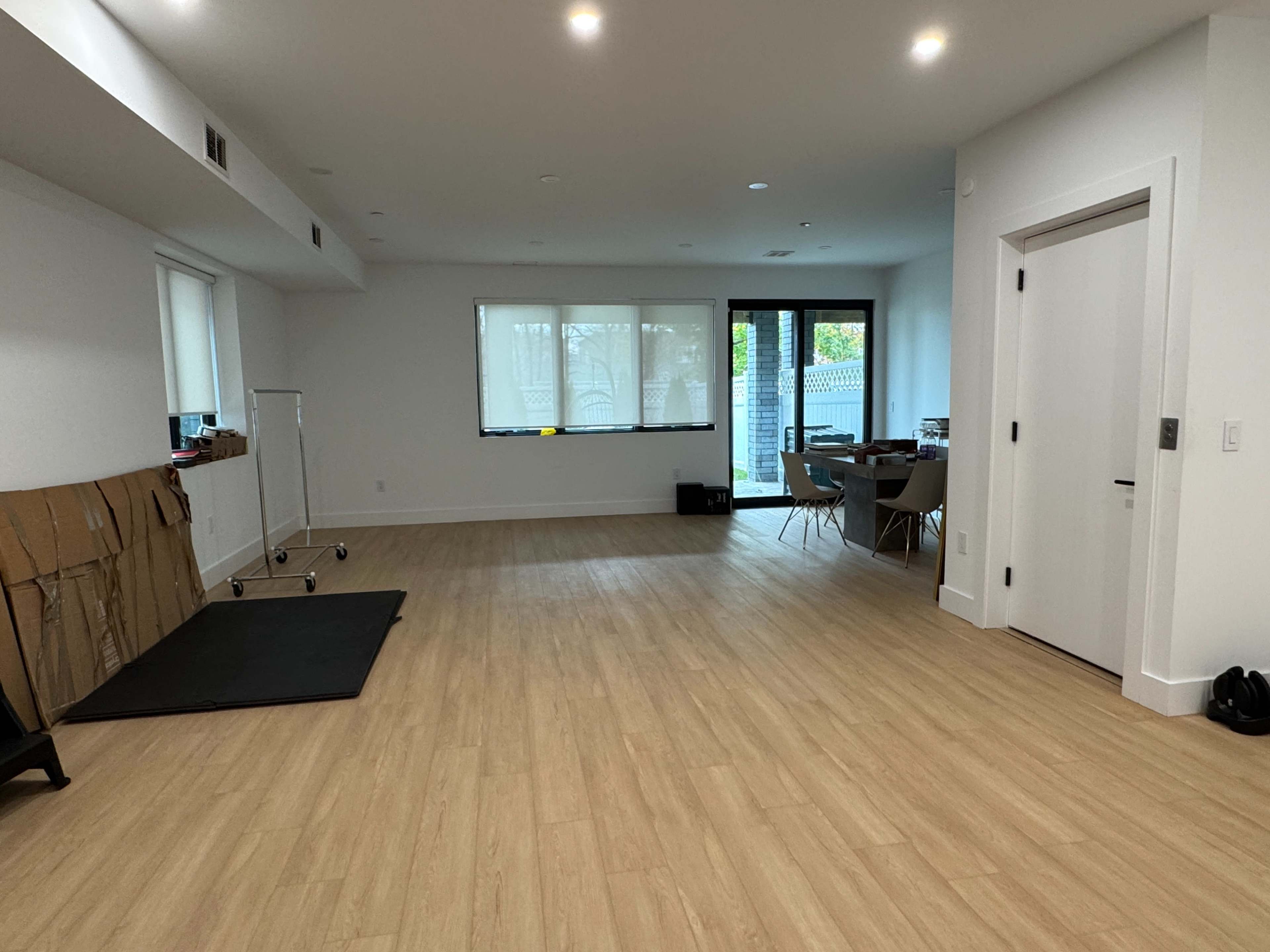 The image shows a spacious, empty room with light wood flooring, a large window, a table with chairs, and a clothing rack against one wall.