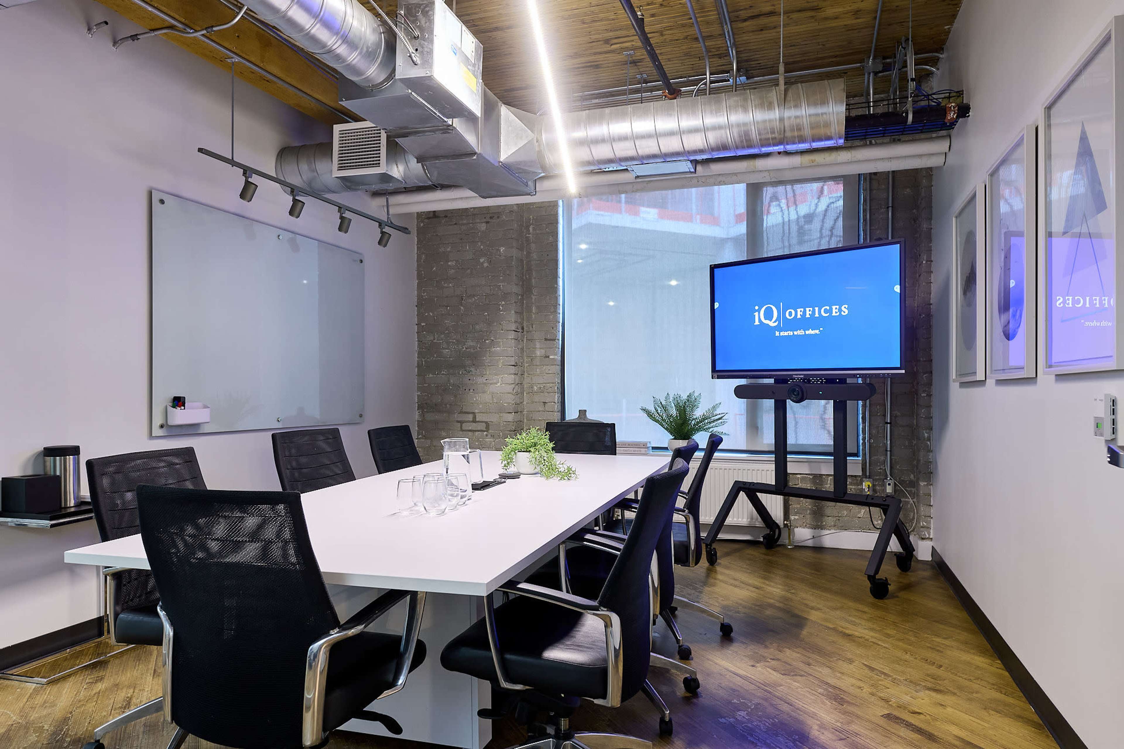 A modern conference room features a long white table surrounded by black mesh chairs, with a large screen on a mobile stand and a glass wall allowing natural light.
