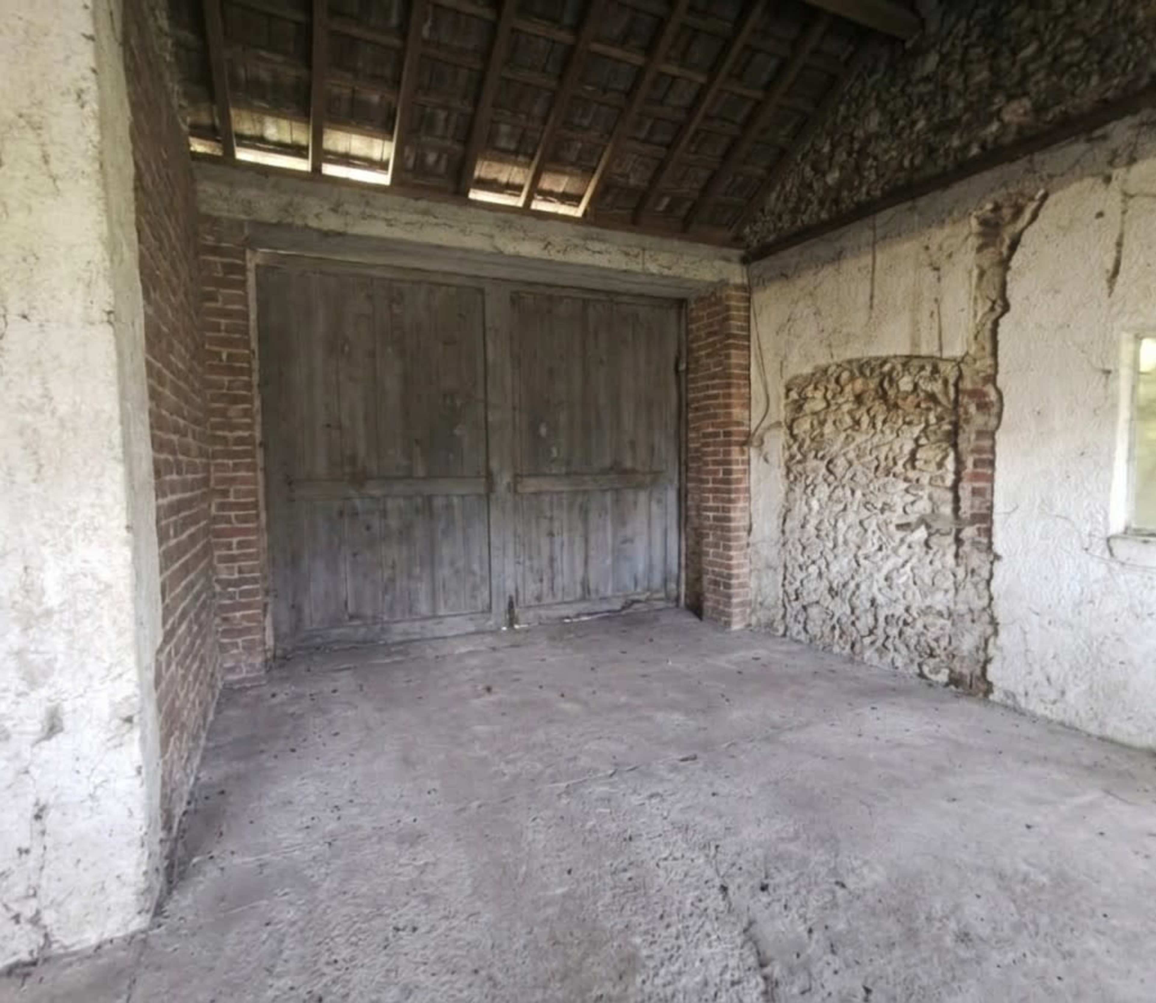 The image shows an empty, rustic interior of a building with wooden double doors and a stone wall.
