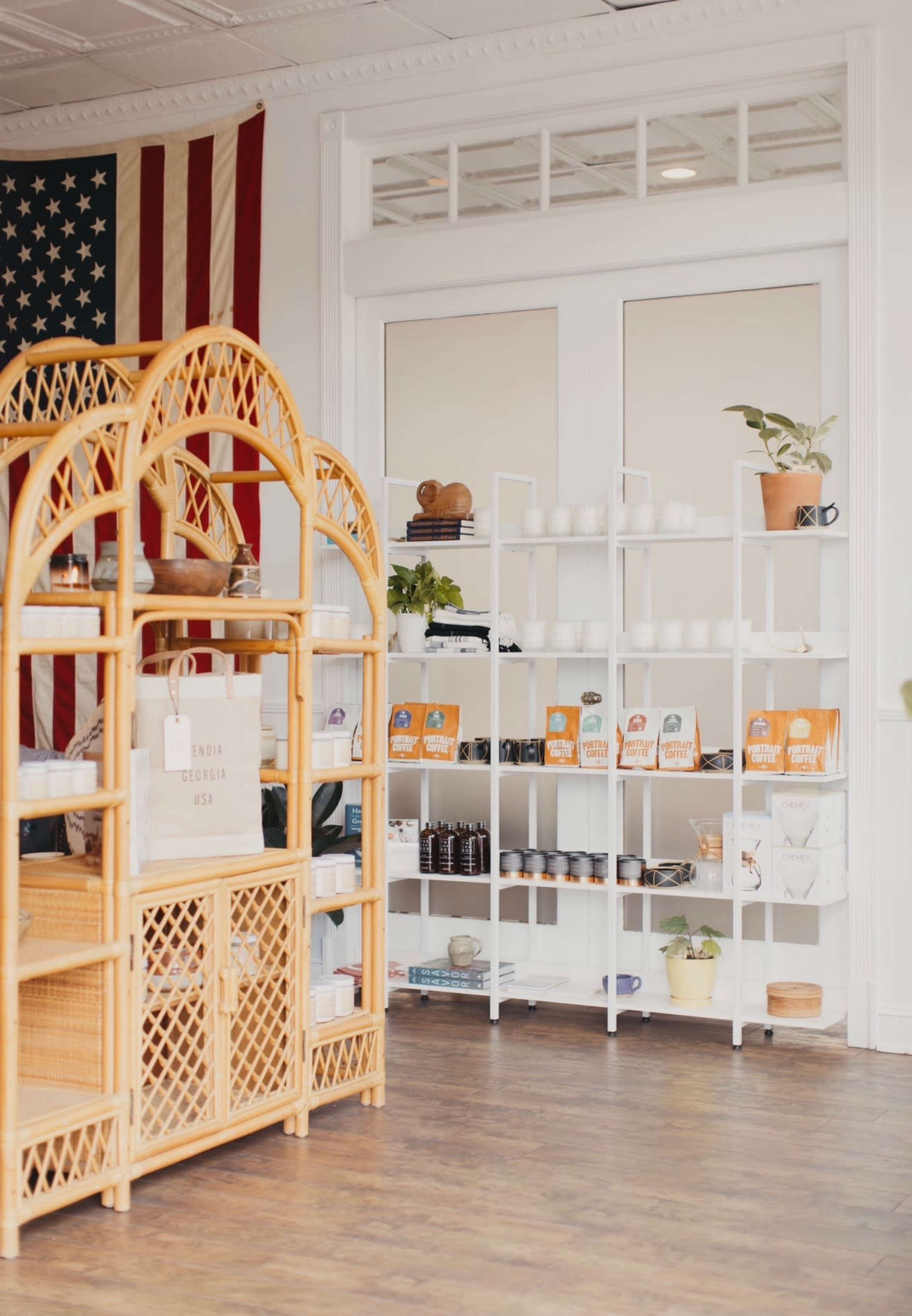 A bright interior features a wooden display shelf with woven accents, an American flag hanging on the wall, and a white shelving unit stocked with products.