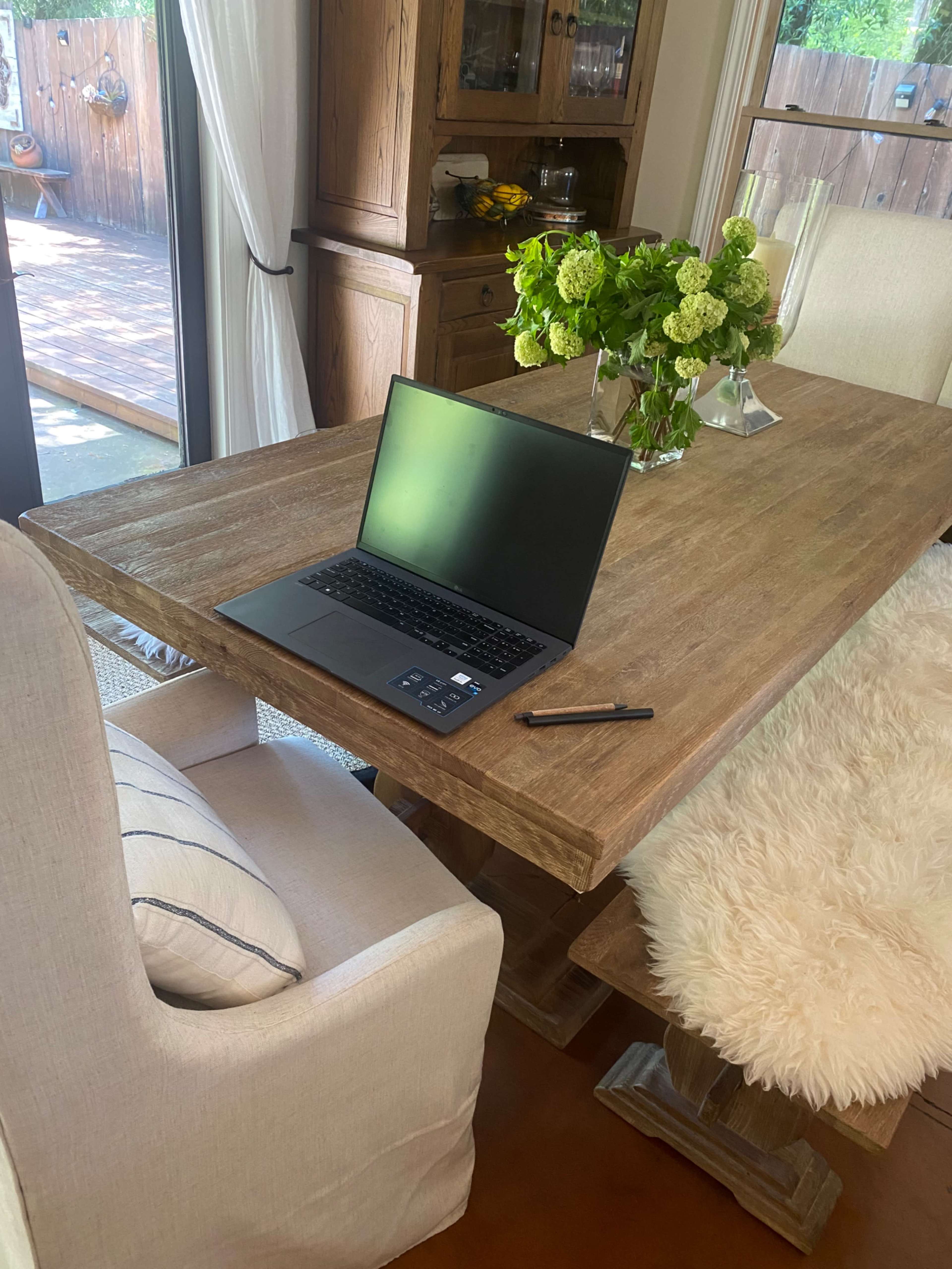 A laptop and a pen are placed on a wooden dining table beside a vase of green flowers.