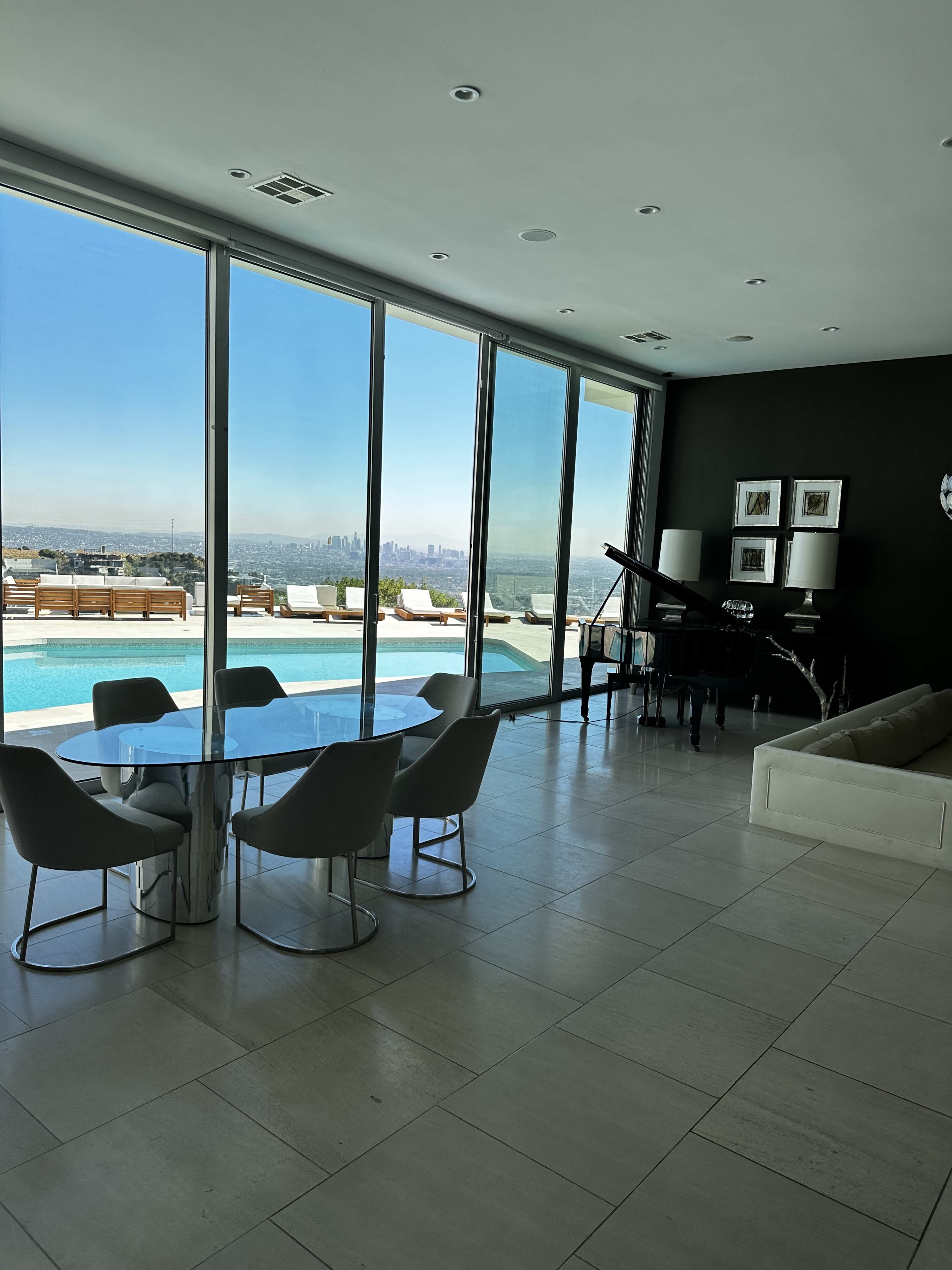 The image shows a modern interior space featuring a round dining table with six chairs, a grand piano, and large windows overlooking a pool and city skyline.
