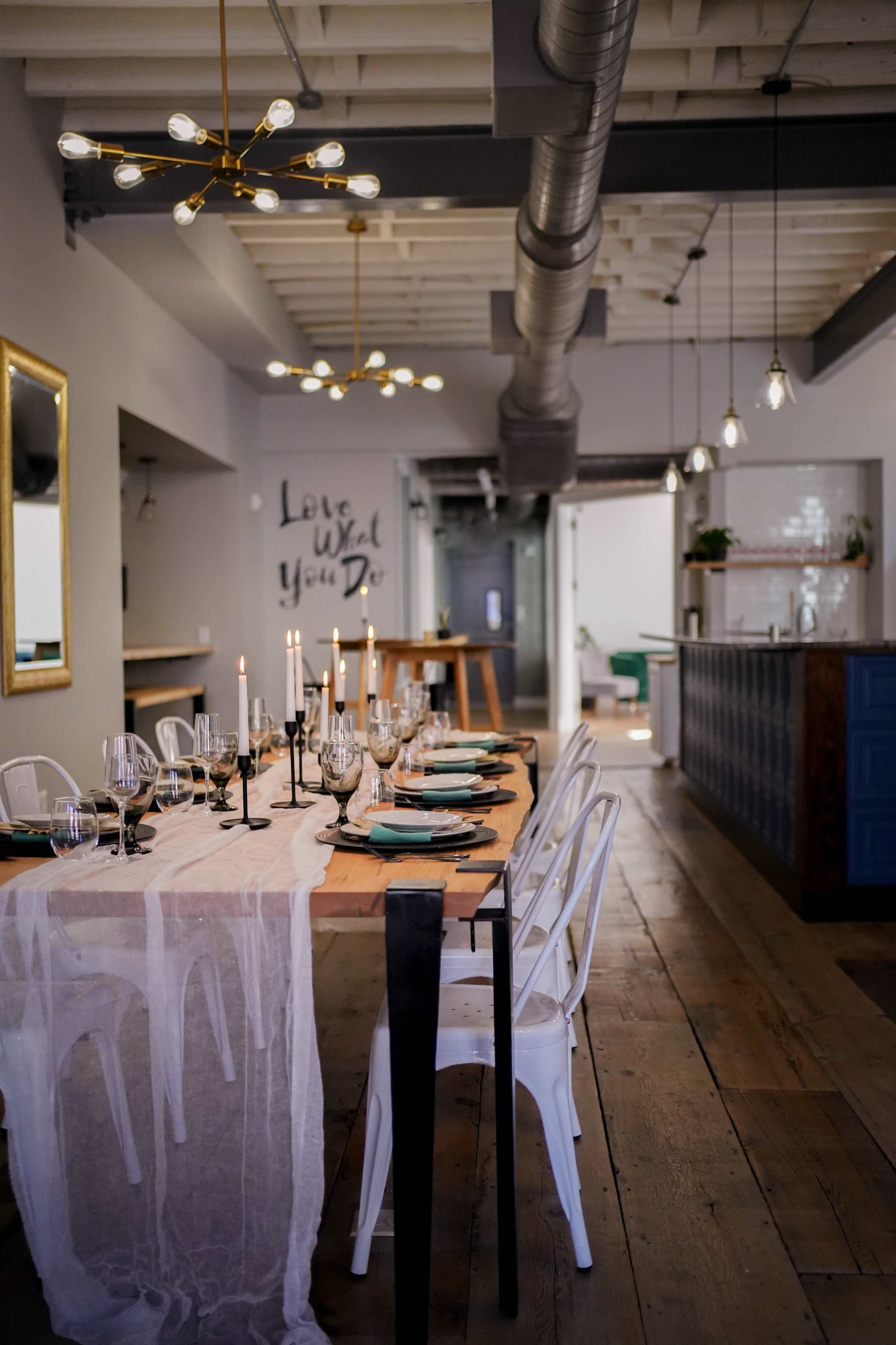 A dining table is set with plates and glasses in a modern interior, featuring a mix of pendant and overhead lighting.