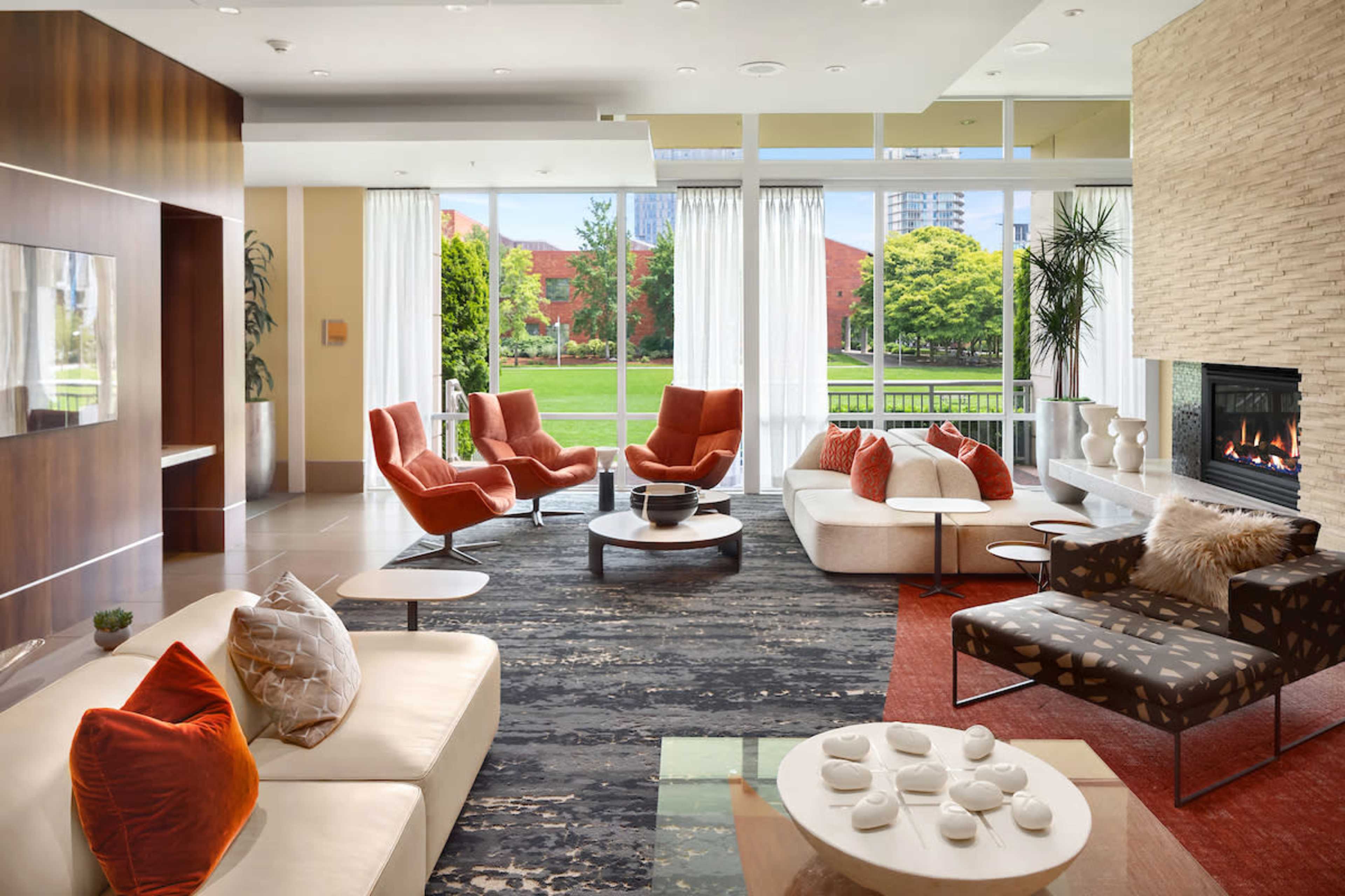 The image shows a modern living room featuring orange-accented furniture, a stone fireplace, and large windows overlooking a green park area.