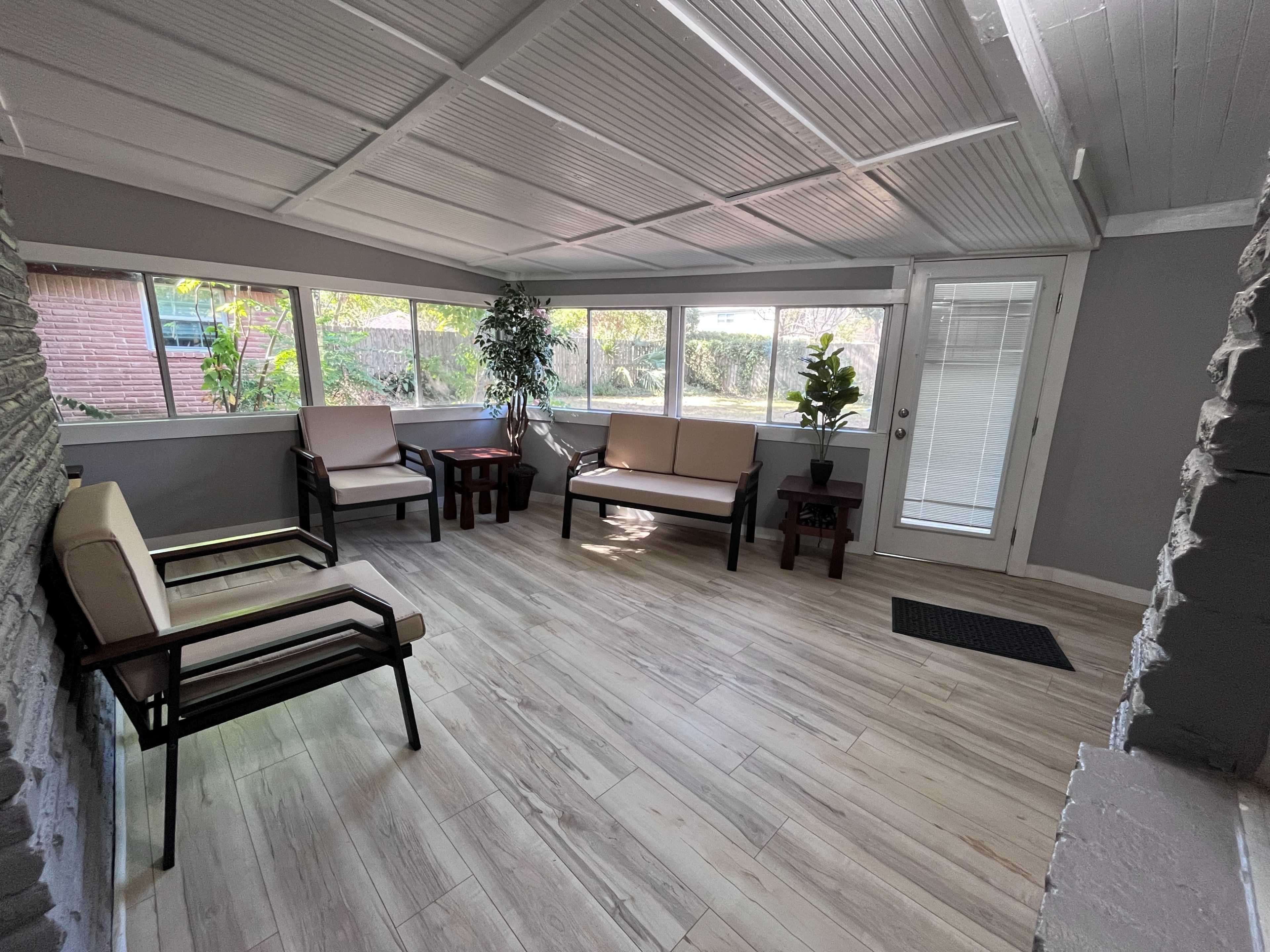 The image shows a sunroom featuring beige furniture, a small wooden table, and large windows that allow natural light to enter the space.