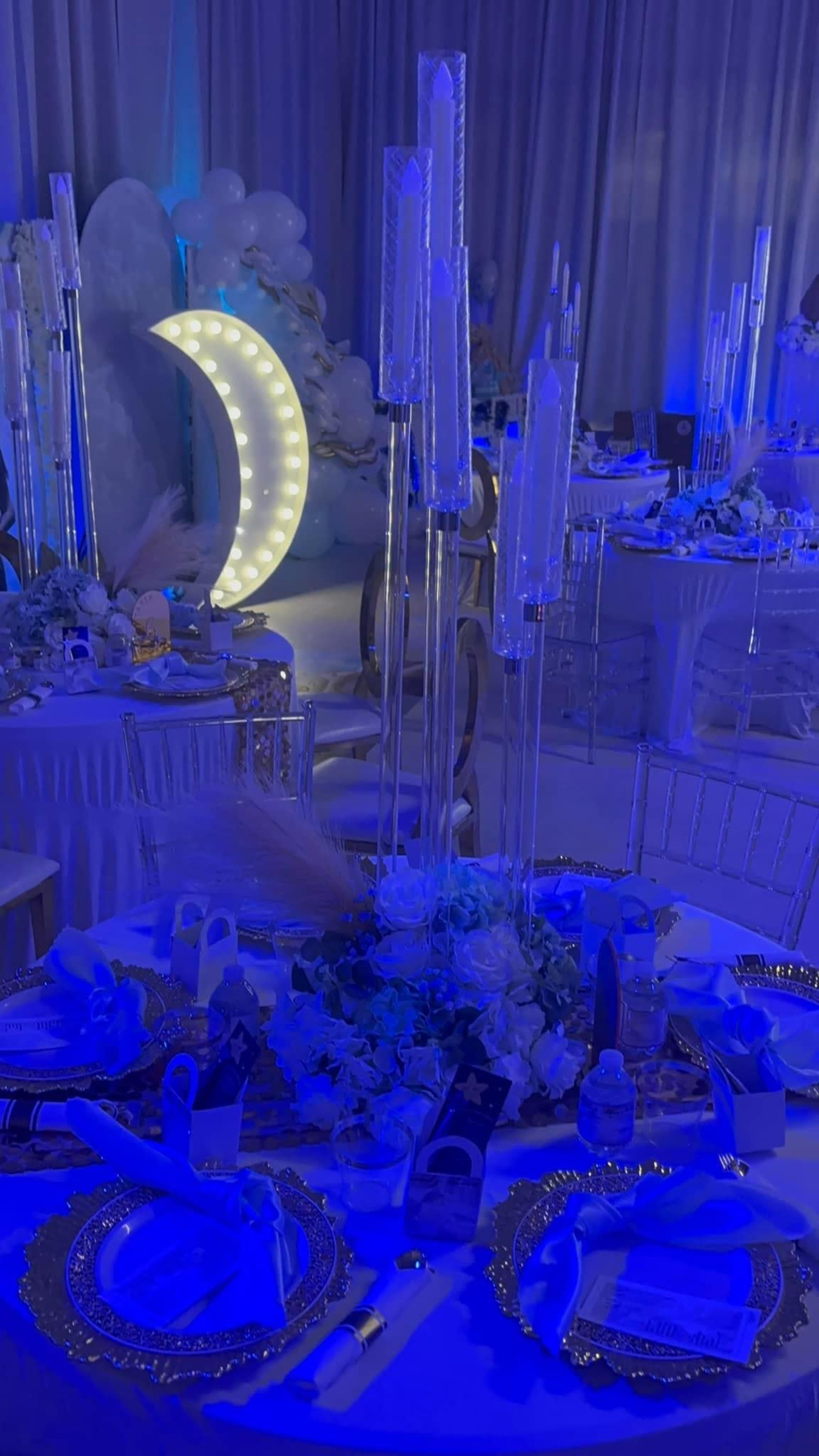 The image shows a beautifully arranged banquet table decorated with blue lighting, elegant tableware, and tall candle holders, with a backdrop featuring a moon and decorative elements.
