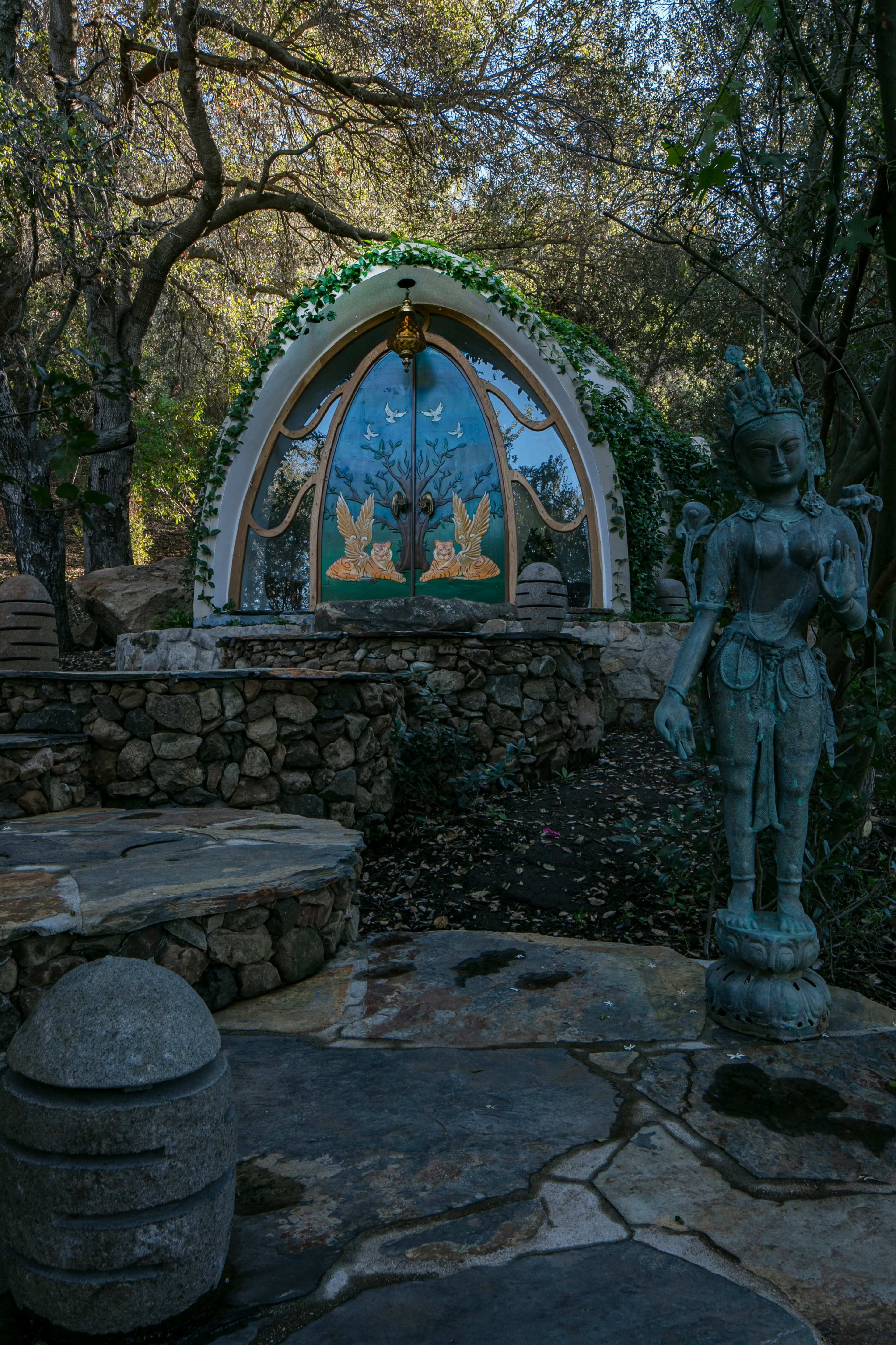 A stone path leads to a decorated entrance featuring a colorful arch with murals and a statue beside it in a wooded setting.