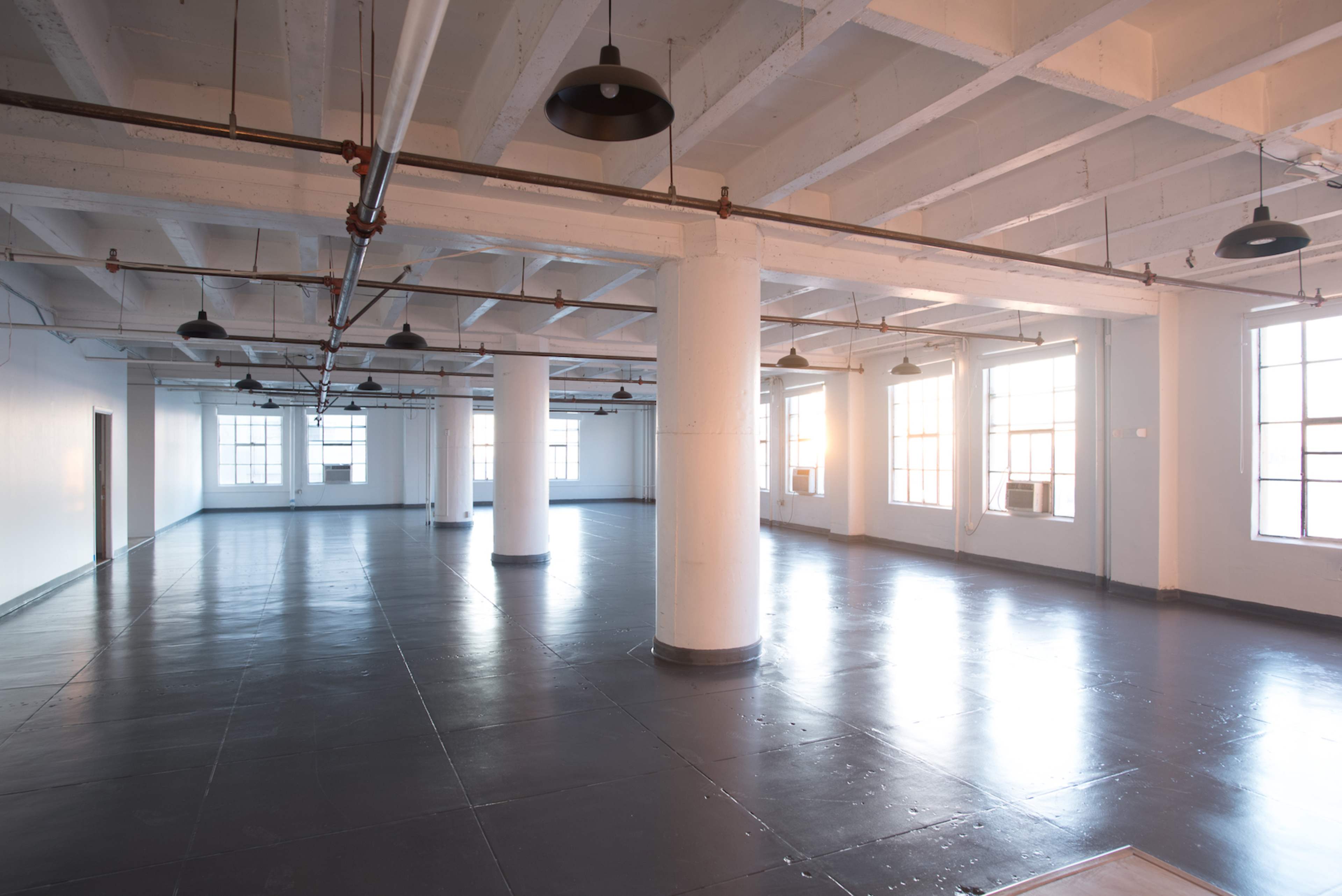 A spacious, empty room with polished dark floors and large windows, illuminated by overhead lights.