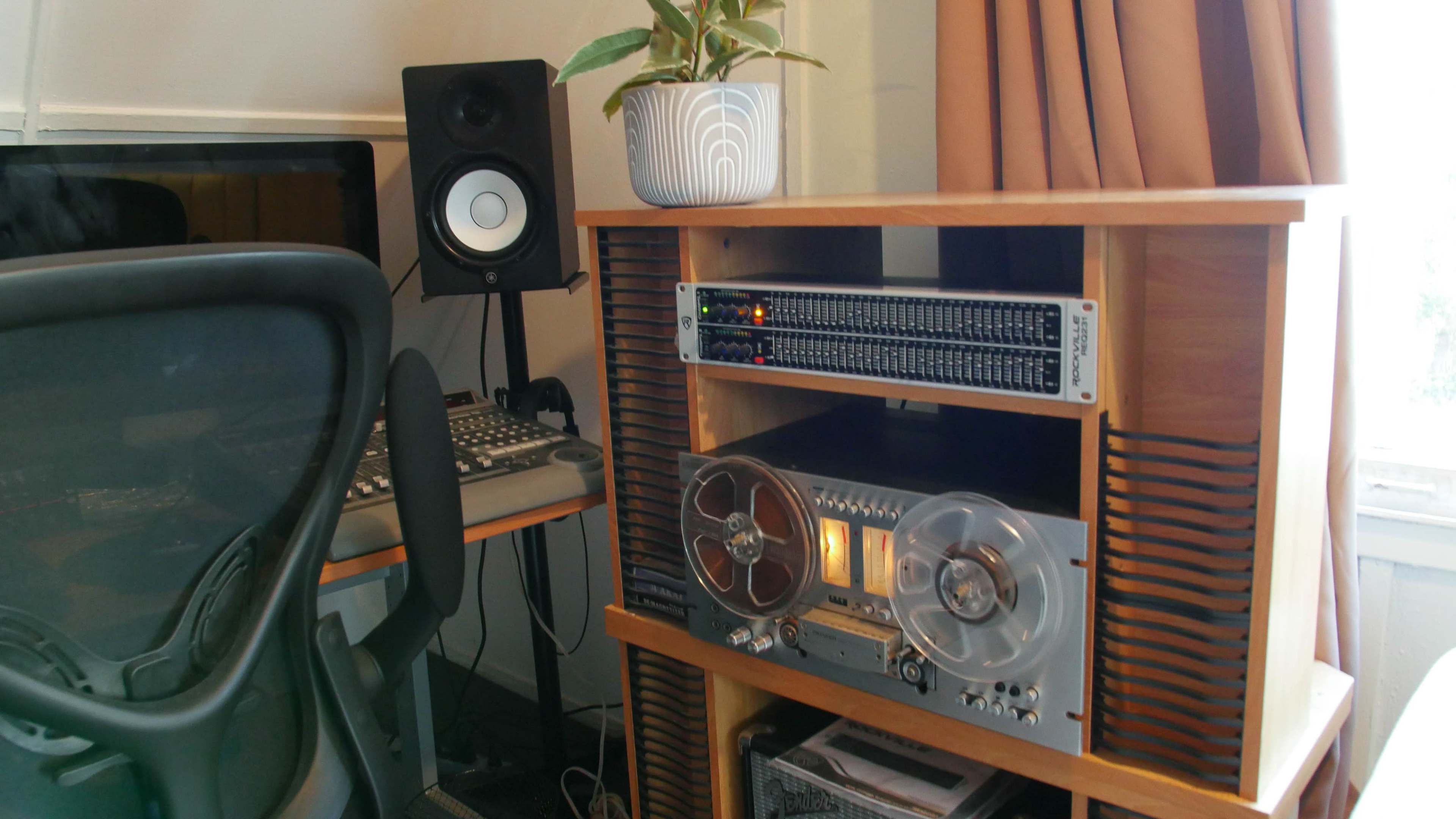 A home workspace with a wooden cabinet containing a vintage reel-to-reel tape recorder, an audio interface, and a potted plant, alongside a computer setup featuring speakers and a mixing console.