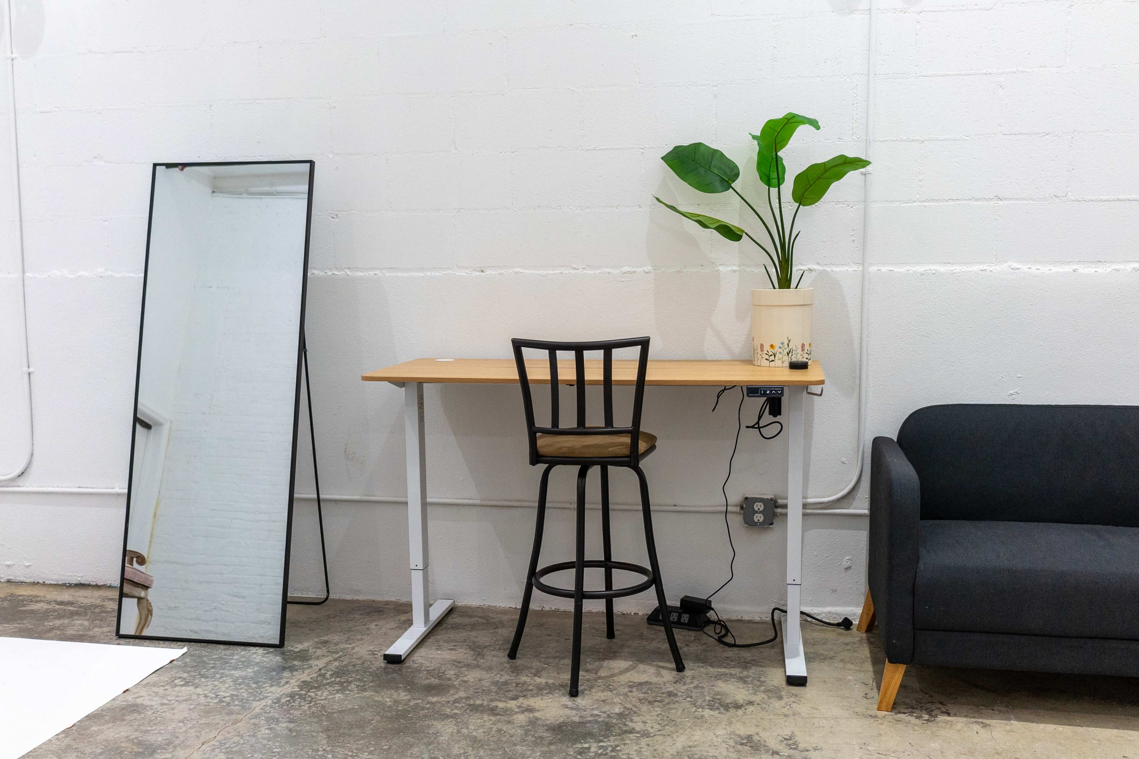 A minimalist workspace features a wooden desk with a high stool, a potted plant, a large mirror, and a dark gray couch against a white wall.