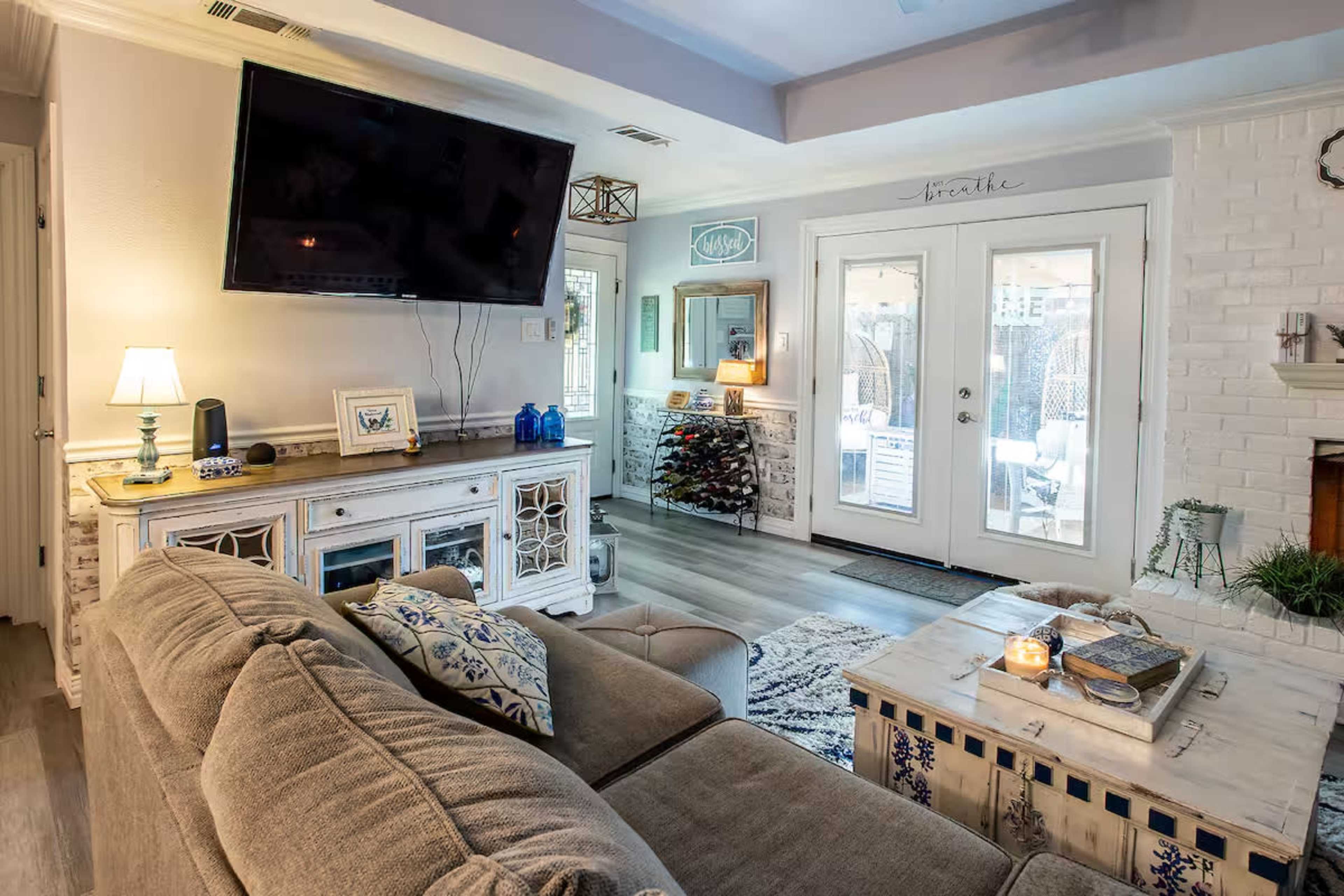 The image shows a cozy living room with a large flat-screen TV mounted on the wall, a comfortable sofa, and a coffee table, with glass doors leading to an outdoor area.
