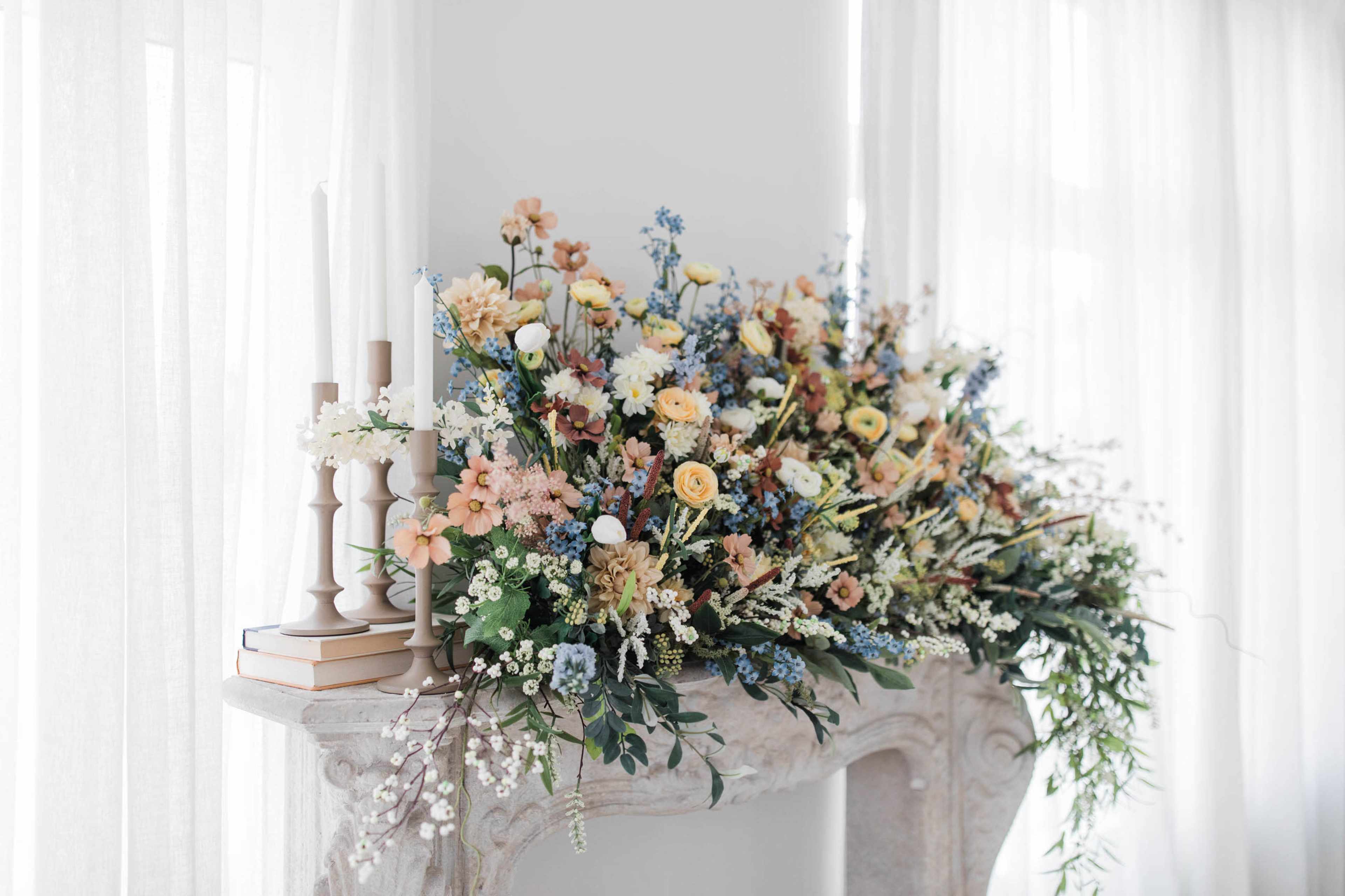 A floral arrangement in pastel colors with various blooms is displayed on a stone mantelpiece, accompanied by several candles and books.