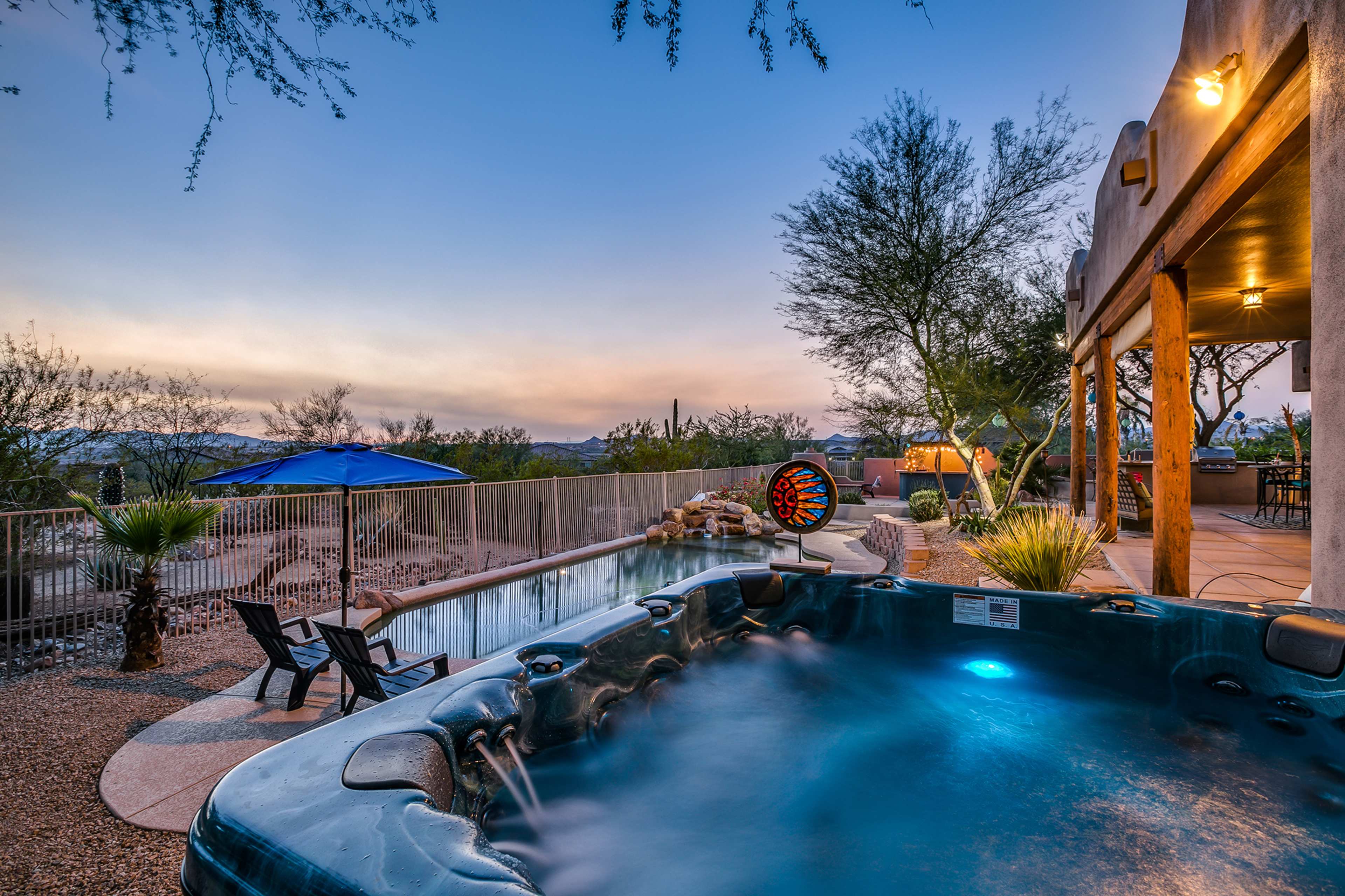 The image shows a hot tub beside a swimming pool, surrounded by desert landscaping and outdoor seating, under a twilight sky.