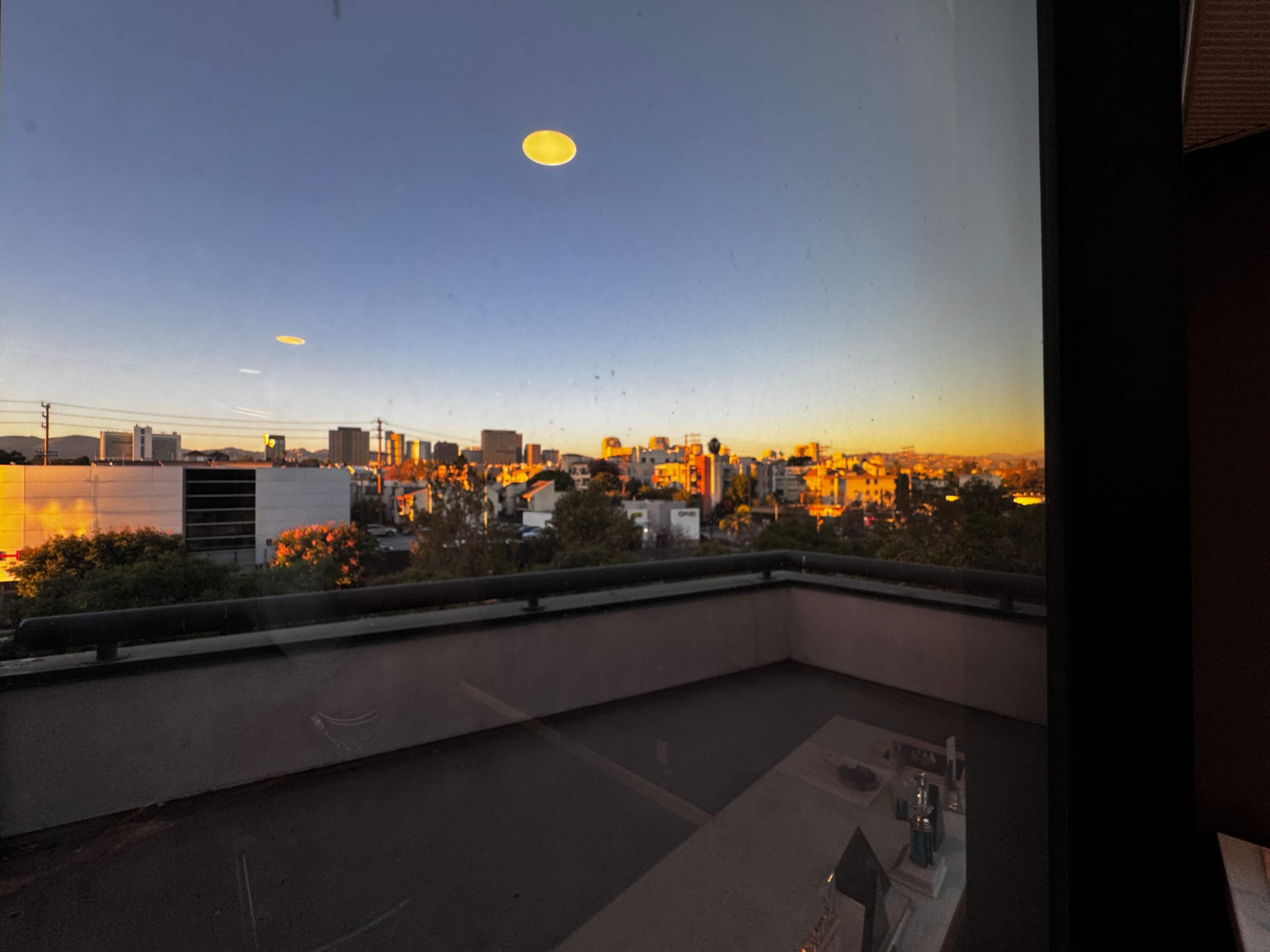 The image shows a city skyline at sunset viewed from a window, with warm light reflecting off the buildings and a balcony in the foreground.