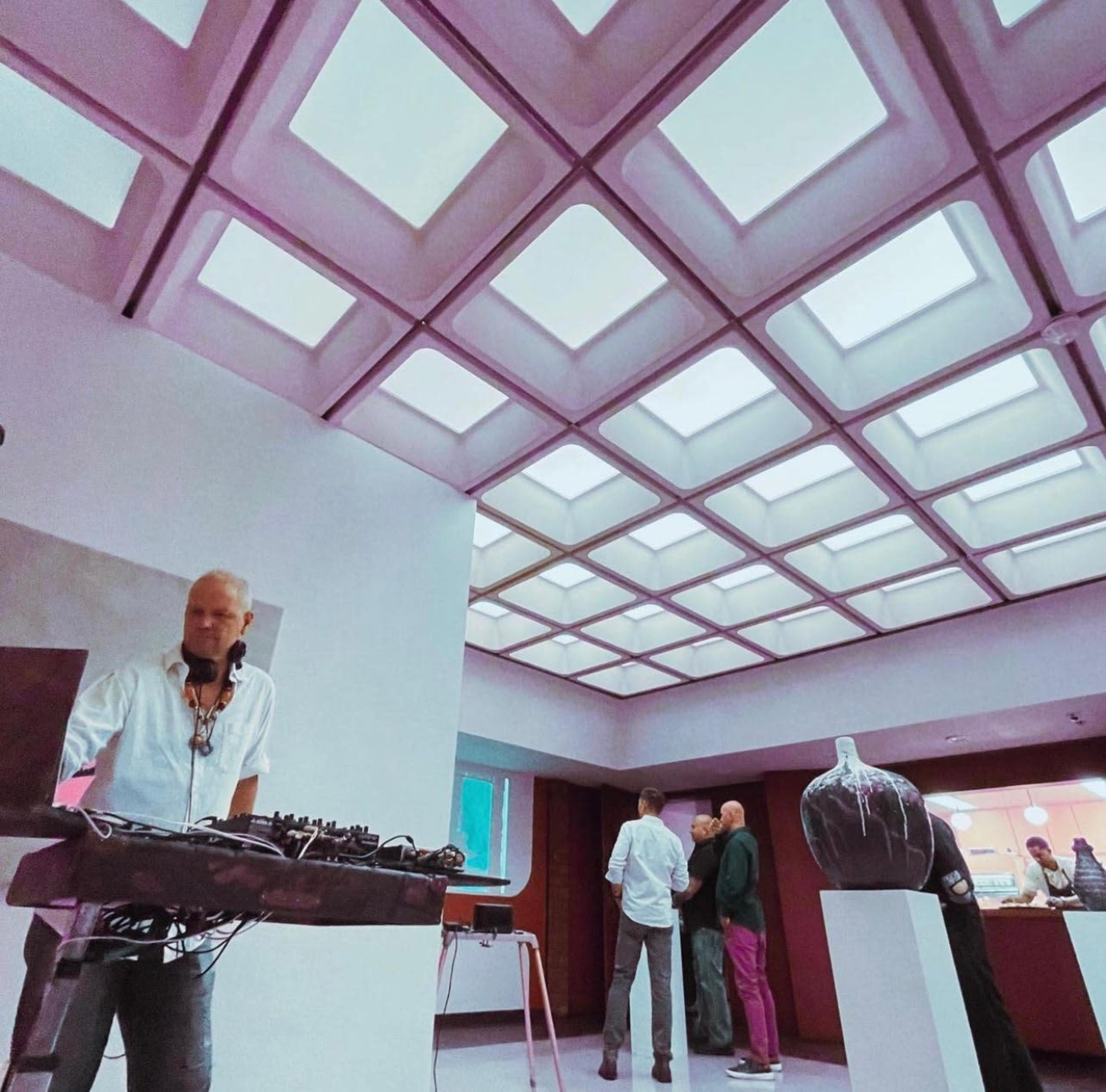 A DJ stands behind a mixing console in an art gallery with a distinctive ceiling and a small group of people in the background.