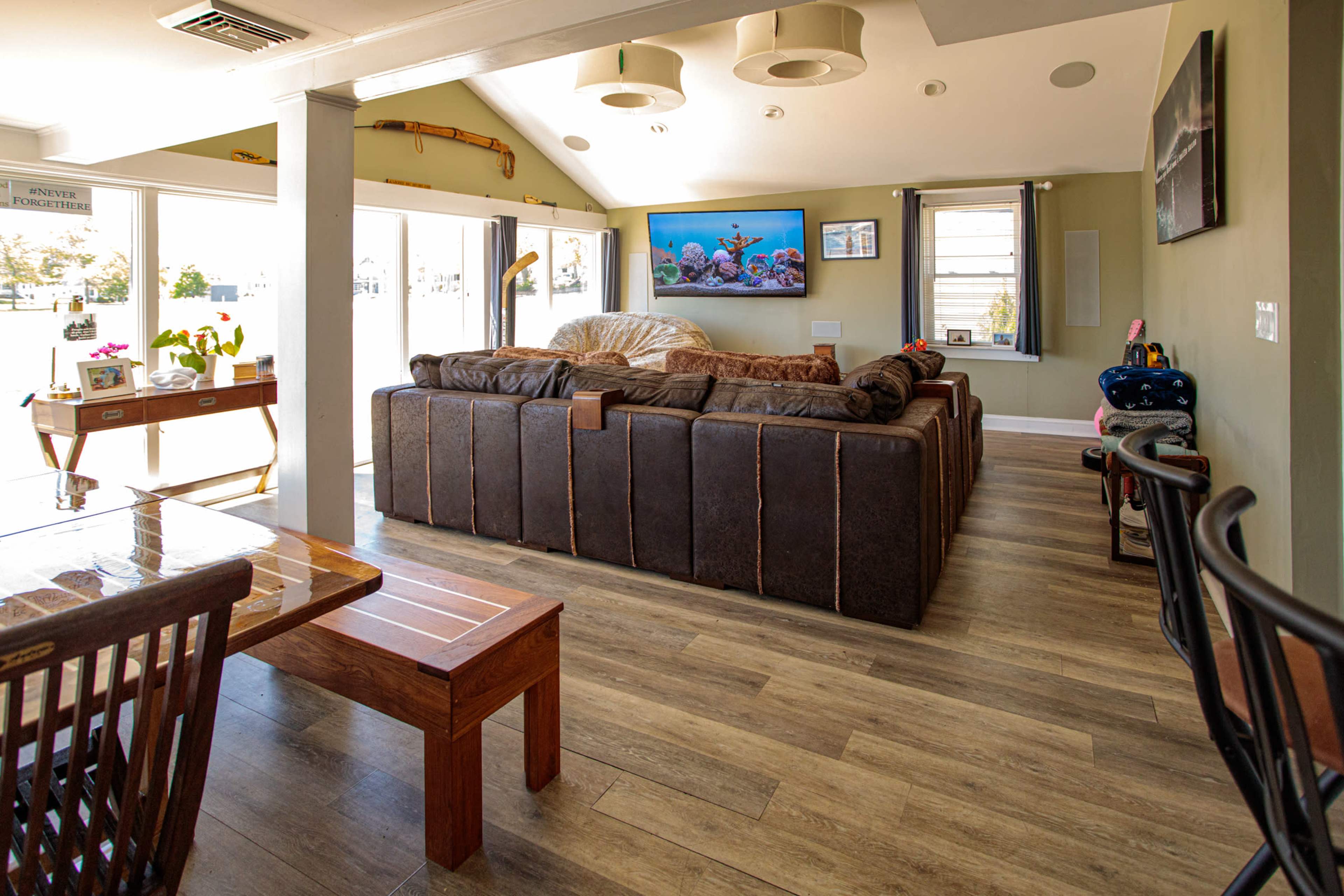 The image shows a spacious living area with a large brown sectional sofa, a dining table, and a flat-screen TV mounted on the wall.