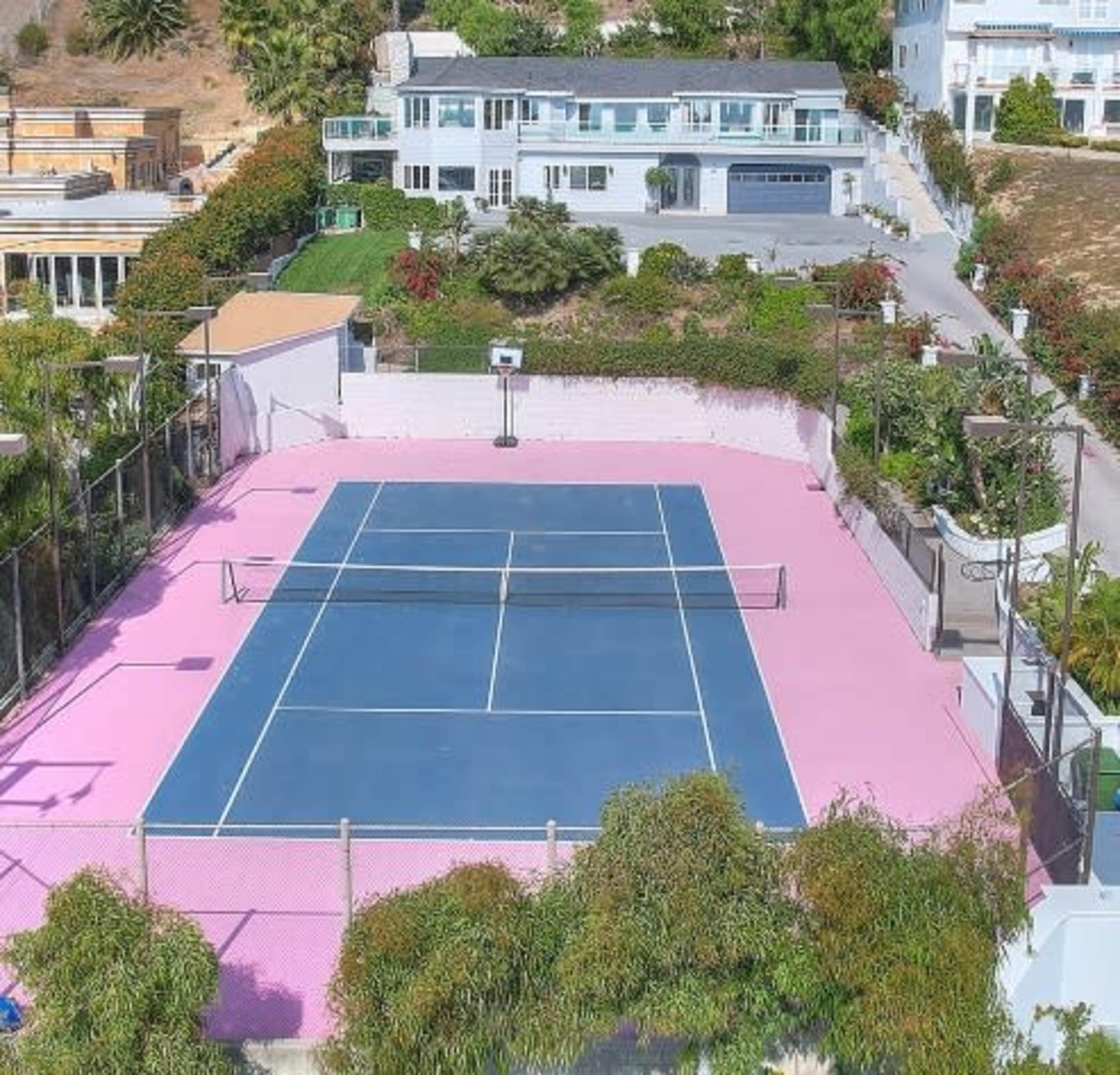 A tennis court with a blue surface and pink surroundings, located in a hillside residential area.