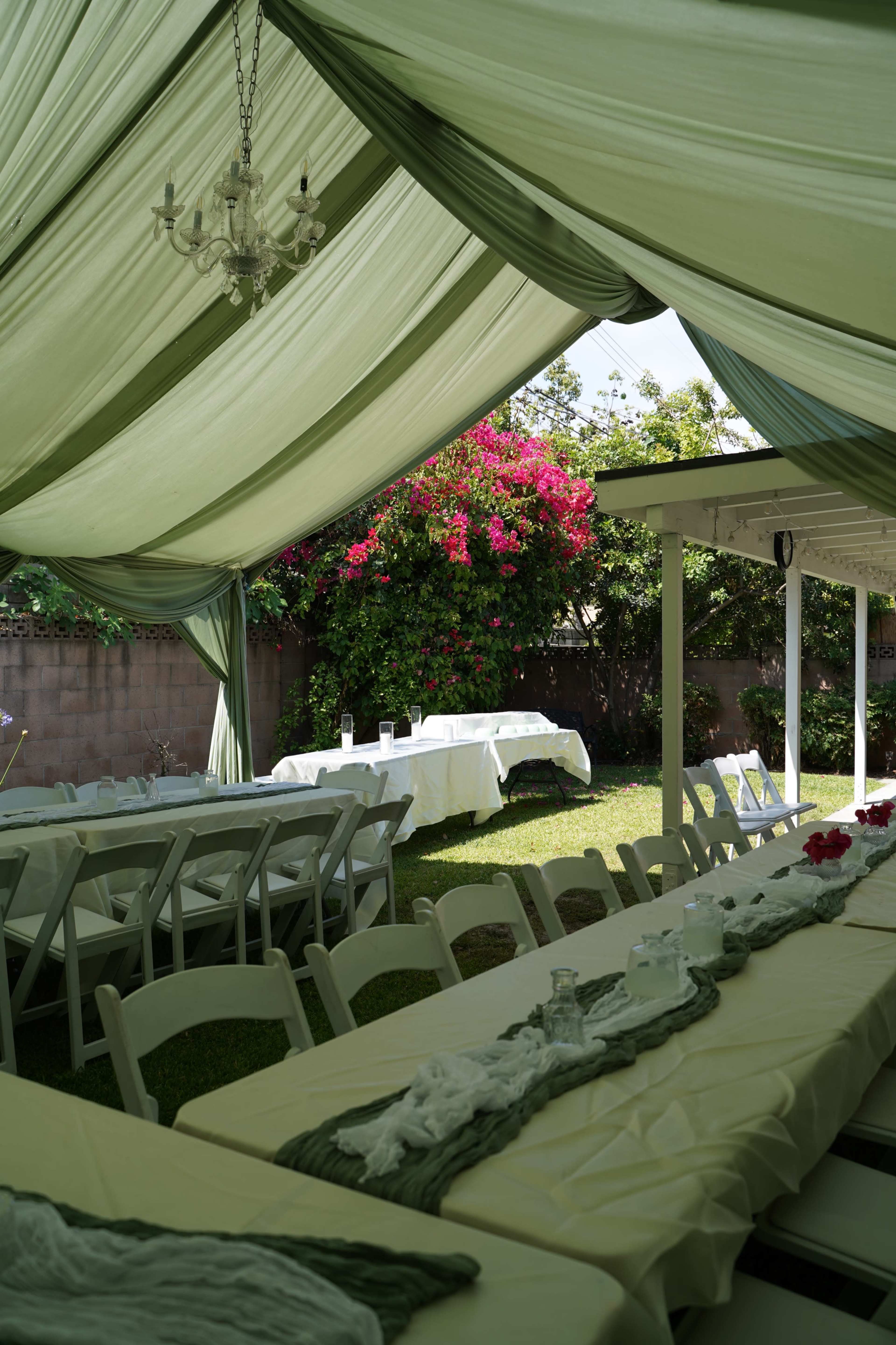 A green and white tent with a chandelier is set up in a garden, featuring long tables covered with cream tablecloths and a backdrop of blooming bougainvillea.