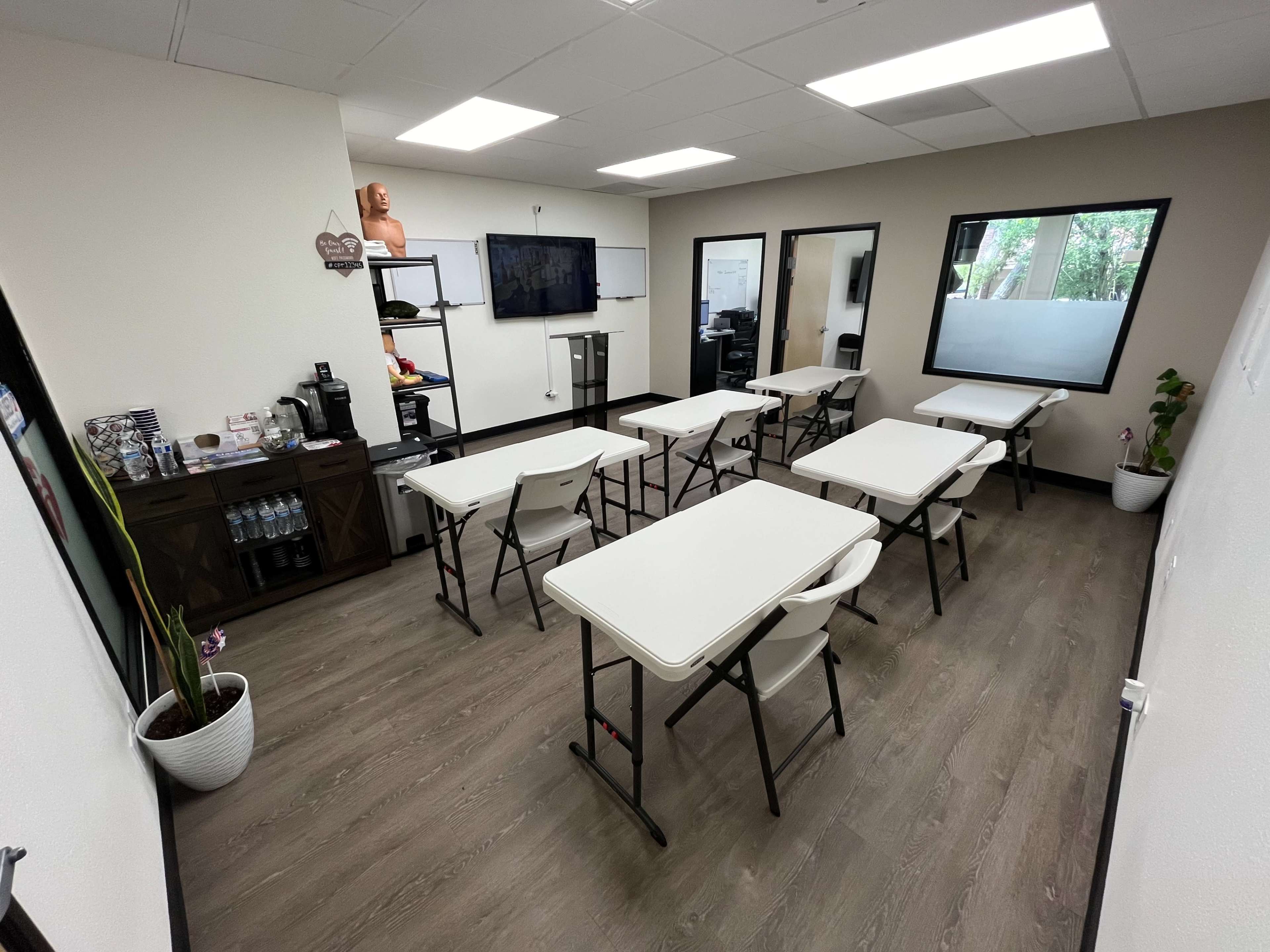 A classroom is set up with several folding tables and chairs arranged for a training session, featuring a television and whiteboard on the wall.