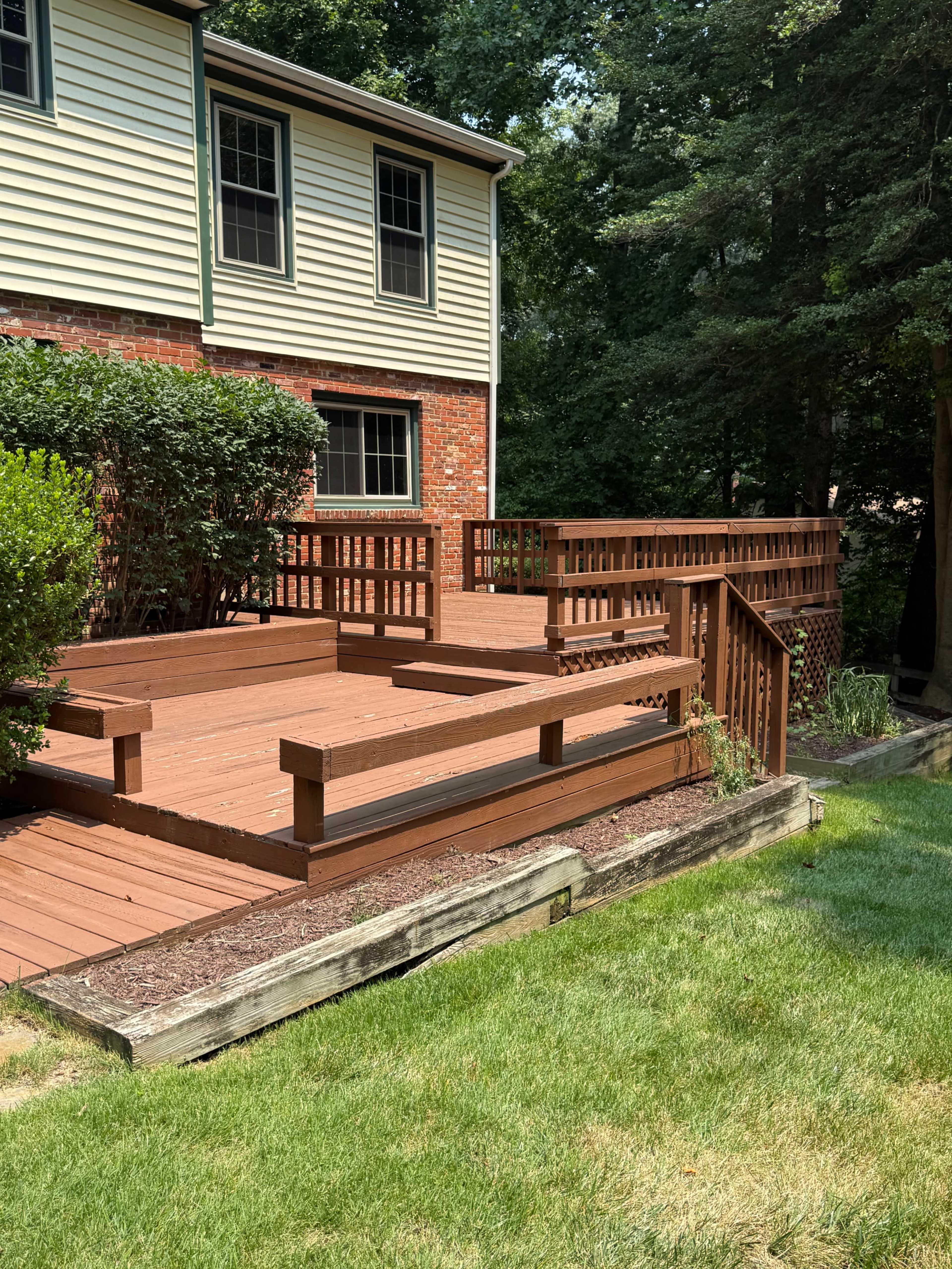 The image shows a wooden deck attached to a two-story brick and siding house, surrounded by trees and a grassy yard.