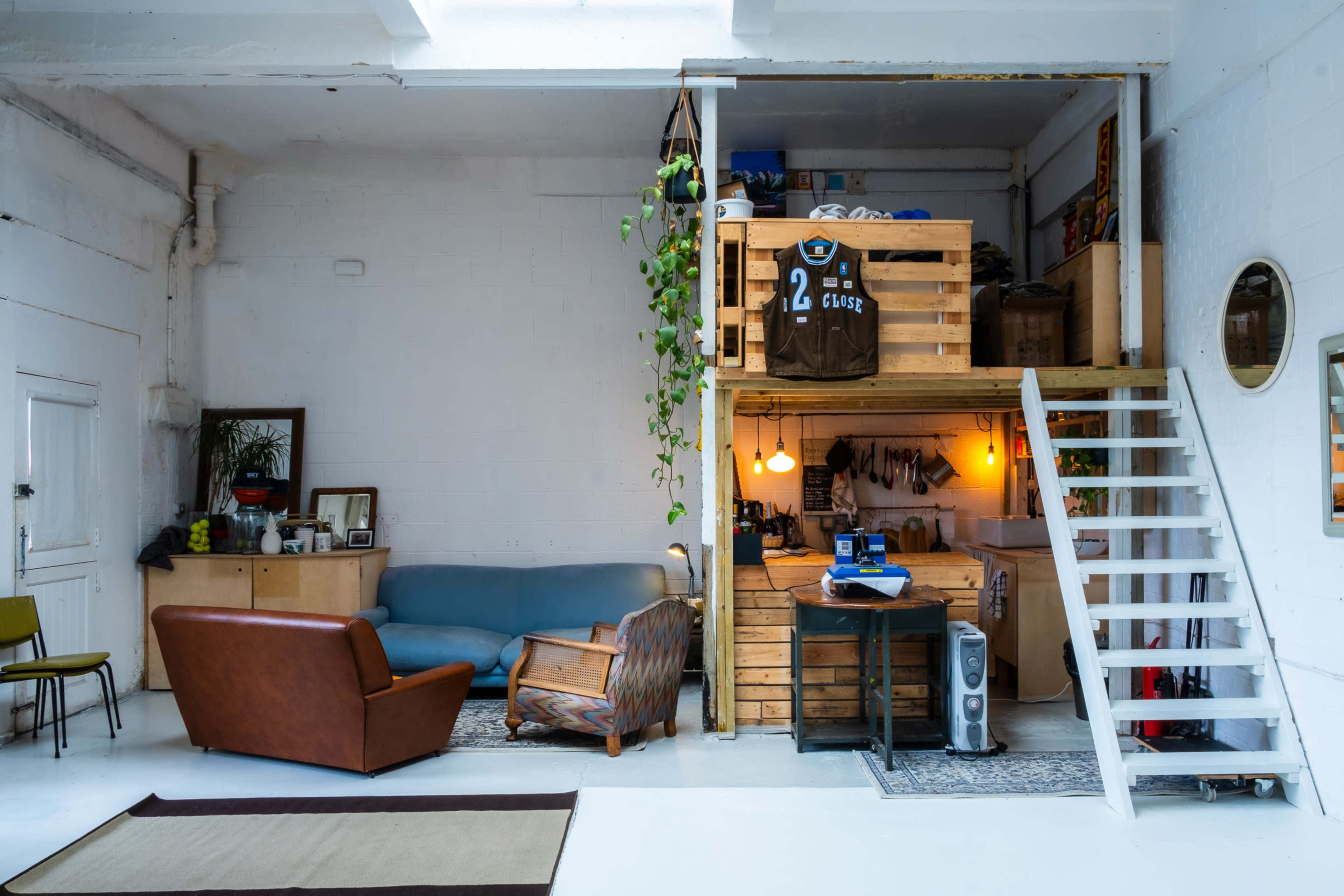 A spacious loft featuring a living area with a blue couch and a brown armchair, alongside a wooden staircase leading to an elevated workspace constructed with pallets.