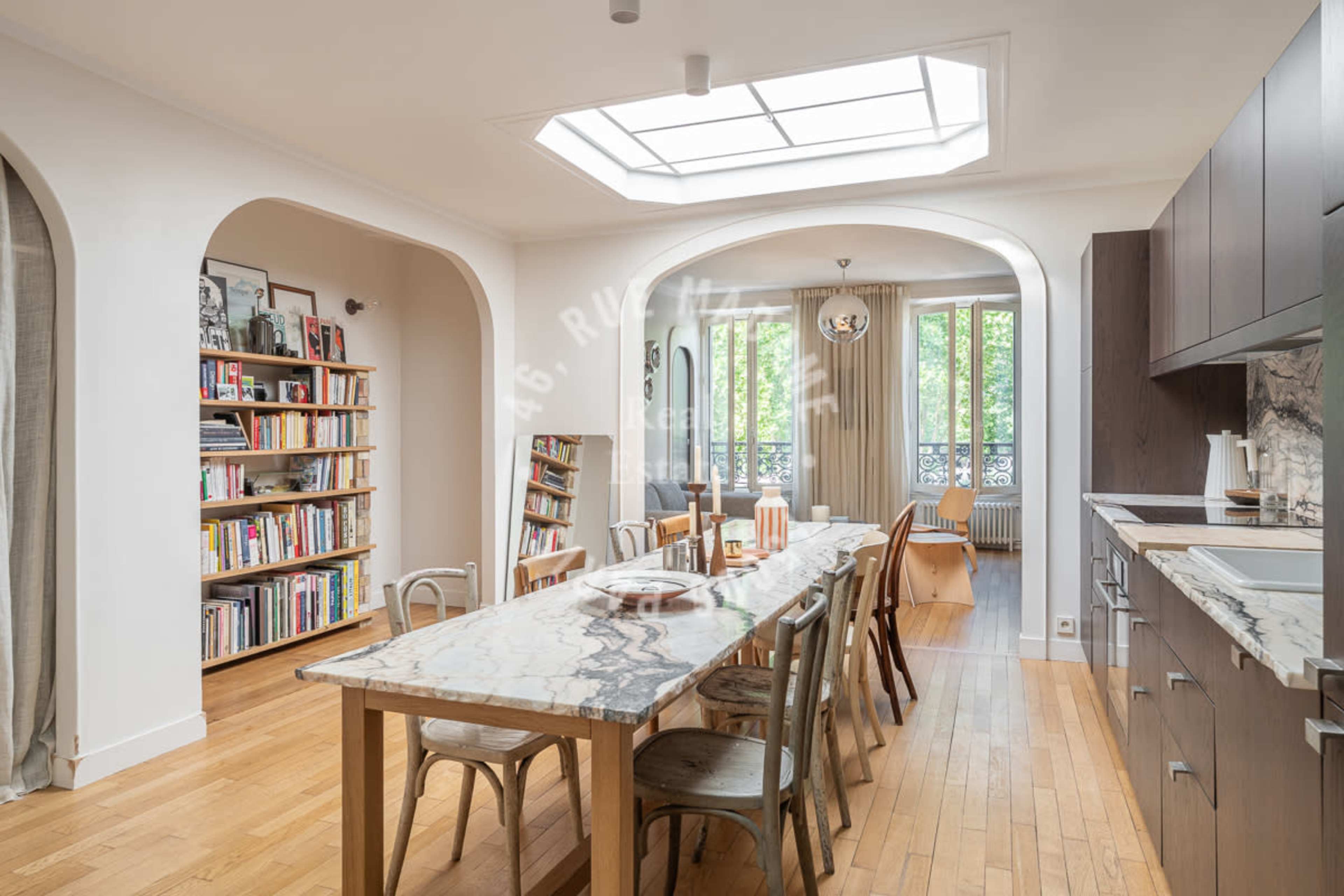 A bright kitchen features a large marble dining table, wooden flooring, and a bookshelf filled with books against a white wall.