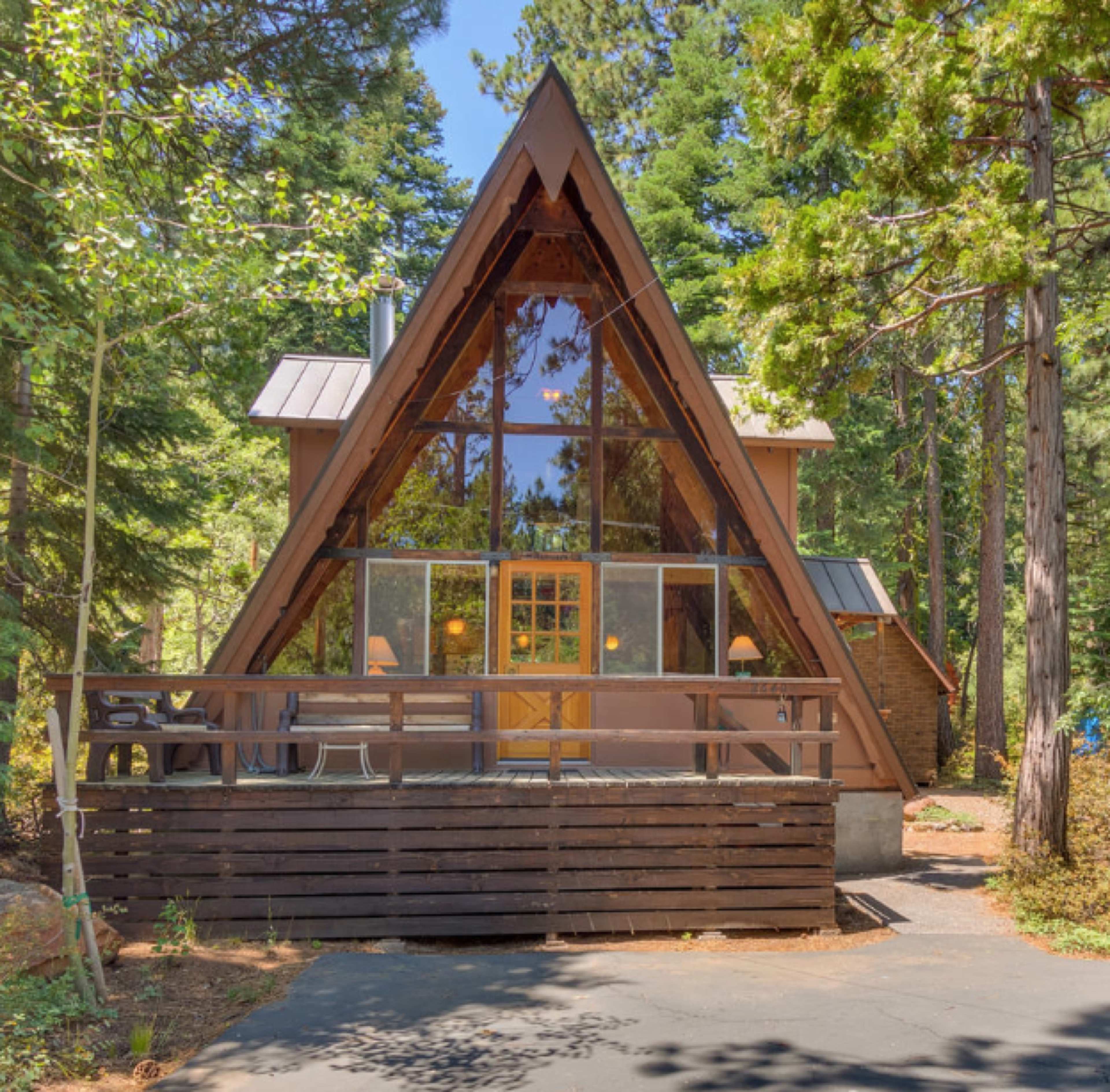 A modern A-frame cabin with large windows sits among tall trees, featuring a wooden deck in the foreground.