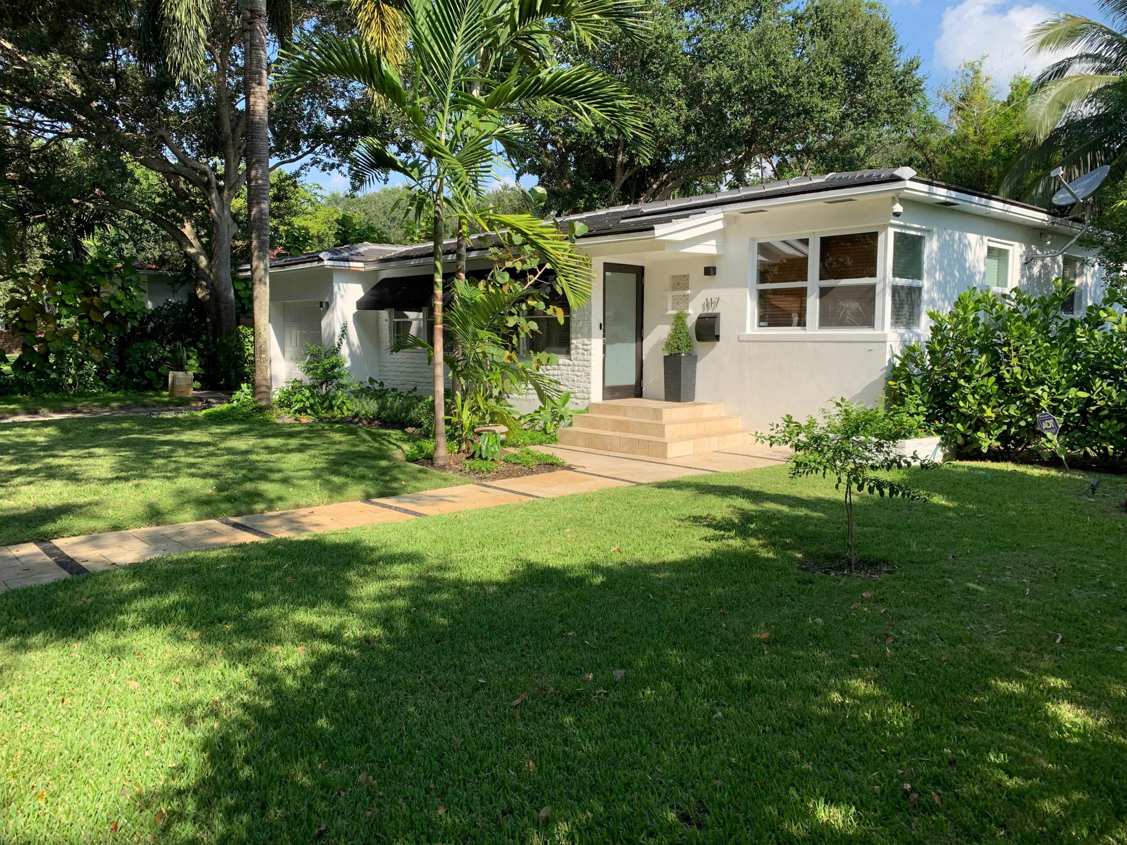 A single-story house with a landscaped yard, featuring a pathway made of stones and surrounded by palm trees and greenery.