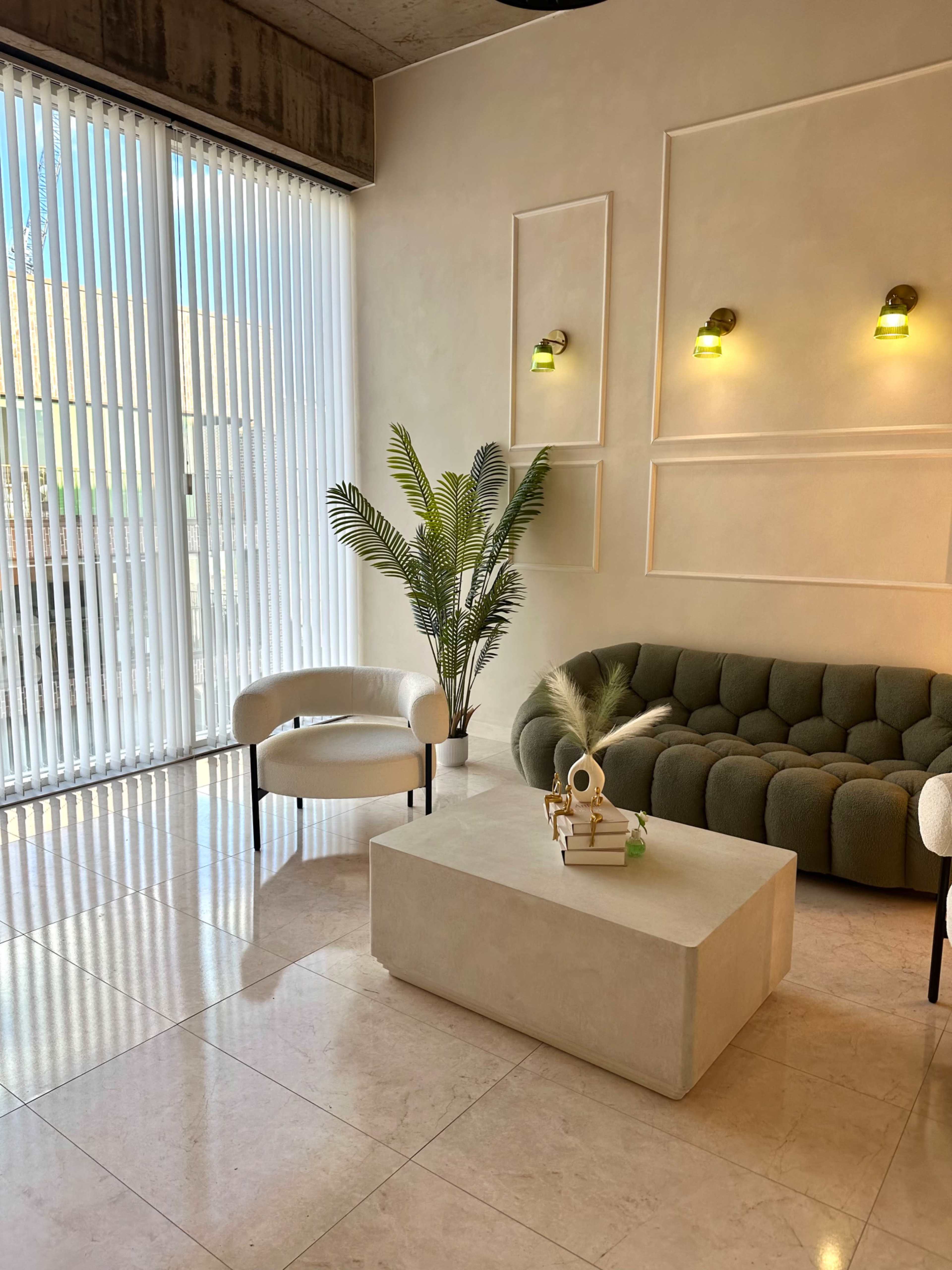The image shows a modern living area featuring a green upholstered sofa, a white circular chair, a stone coffee table, and vertical blinds letting in natural light.