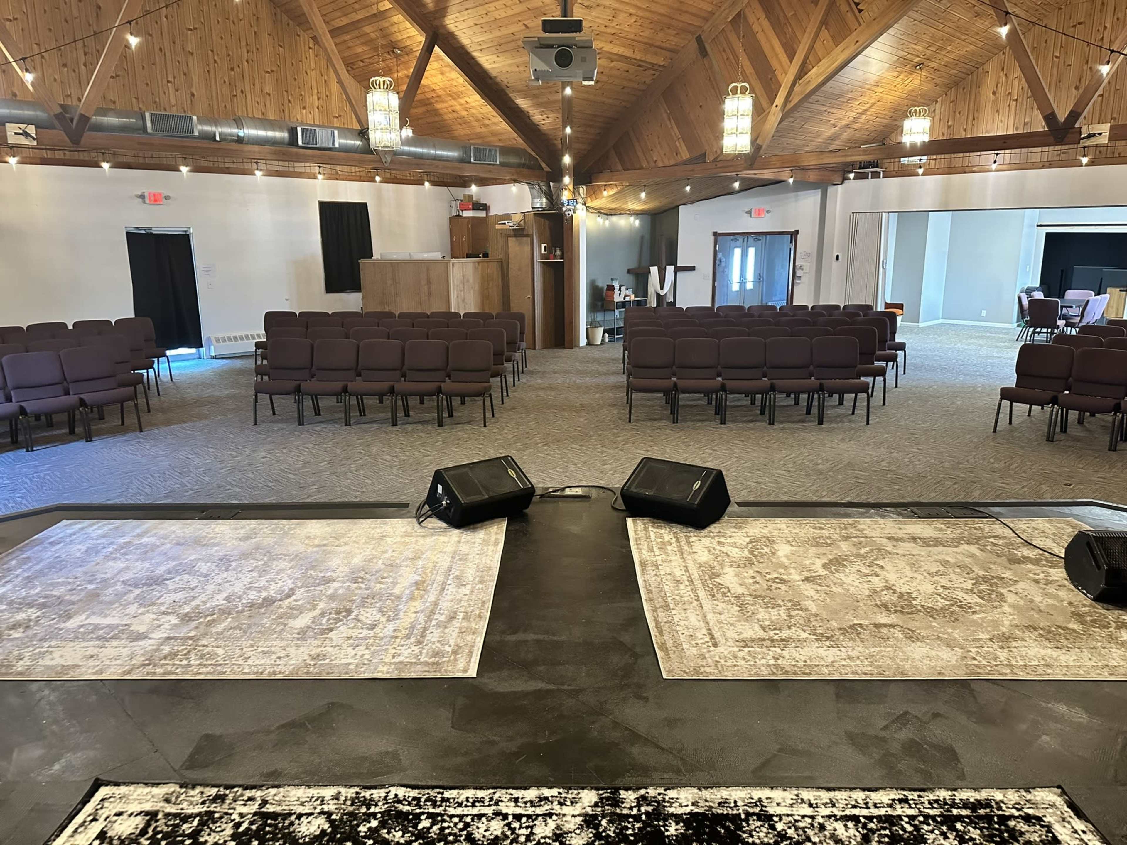 A spacious indoor venue with a raised stage, rows of seating, and wooden beam ceilings.