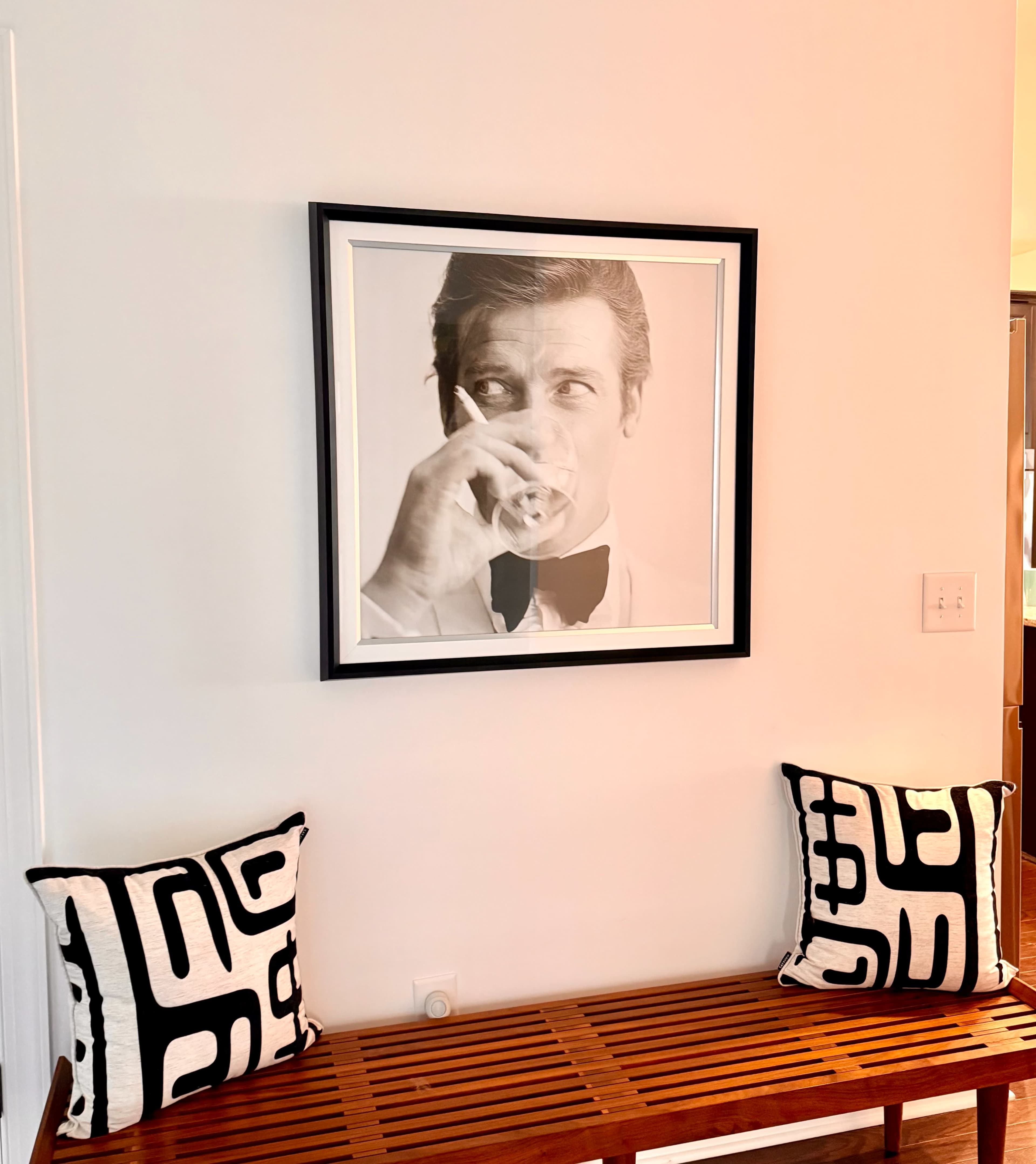 A framed black-and-white portrait of a man sipping a drink is mounted on a white wall above a wooden bench, flanked by two patterned cushions.