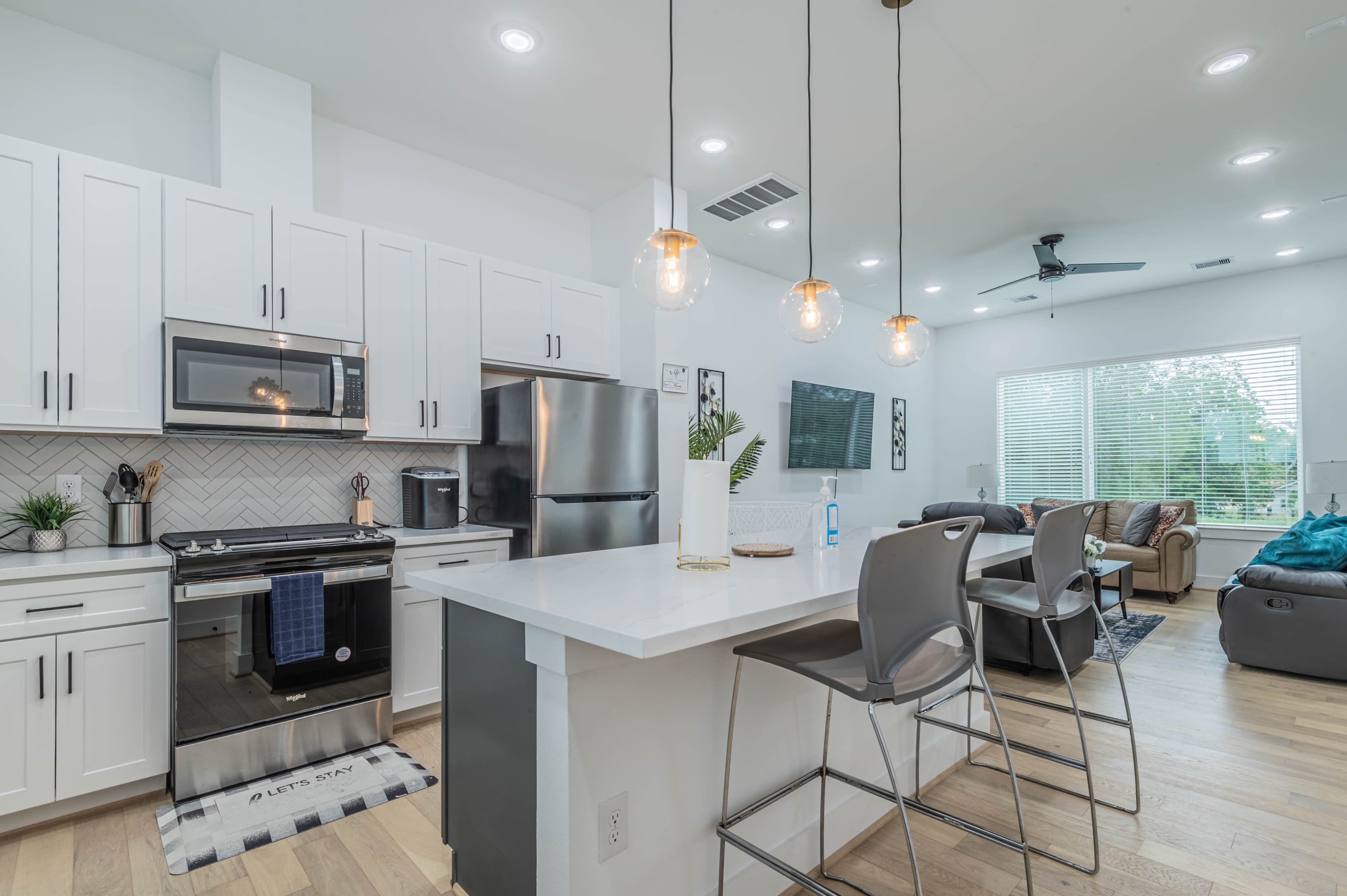 A modern kitchen with white cabinetry, stainless steel appliances, and a stylish countertop that opens into a living area with a large window and comfortable seating.