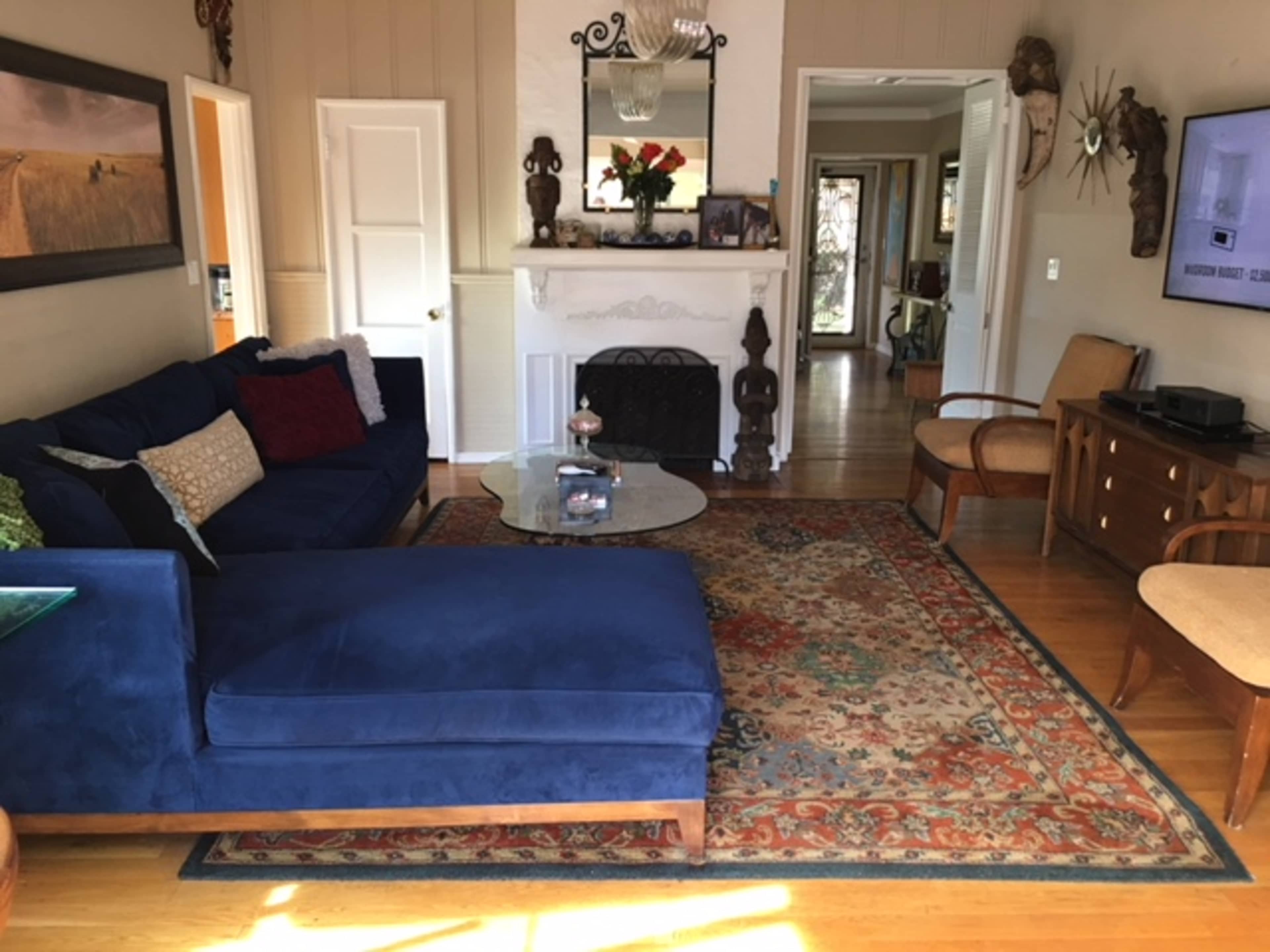The image shows a living room featuring a blue sectional sofa, a glass coffee table, a colorful area rug, and a fireplace adorned with decorative items.
