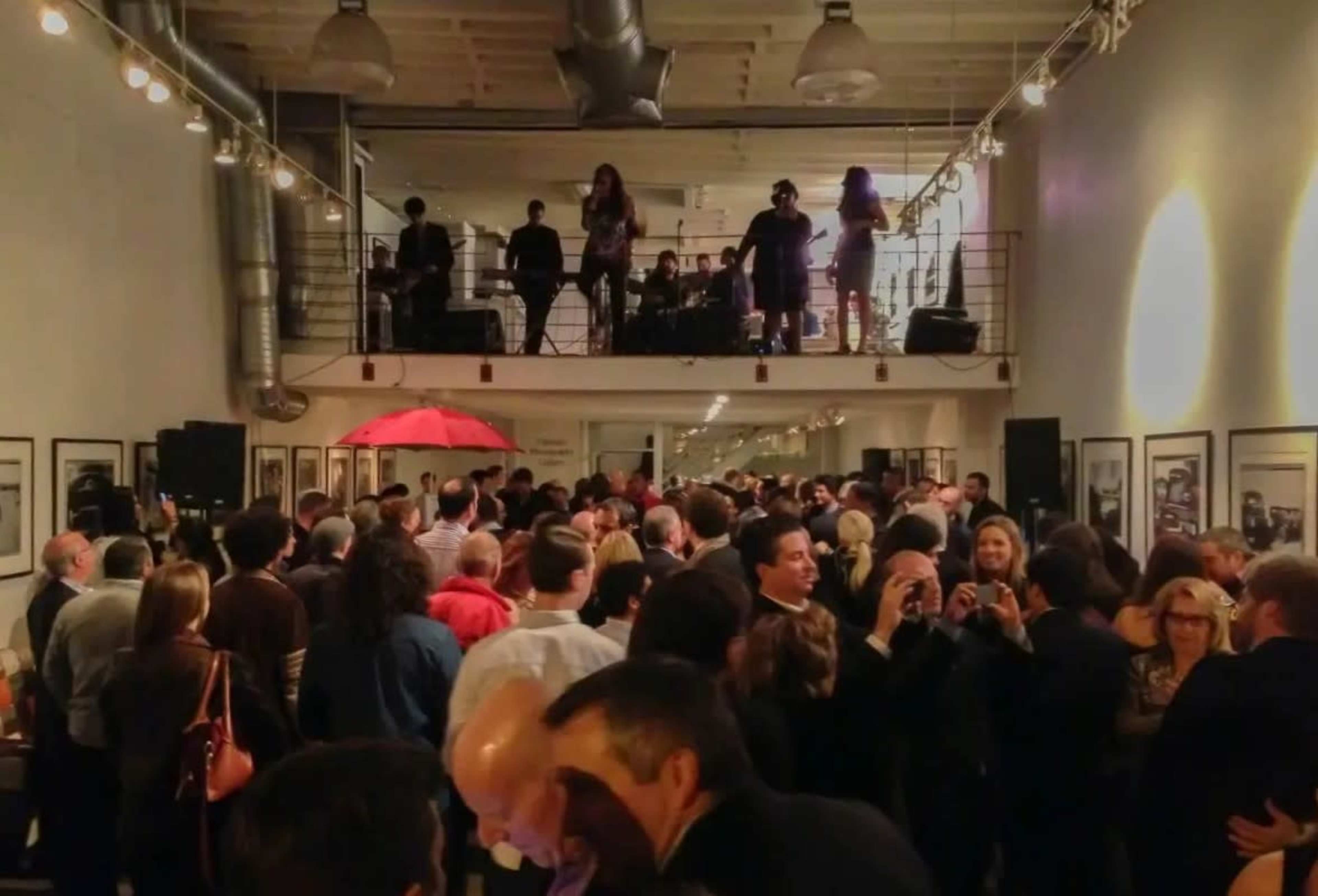 A crowded art gallery features a live band performing on a raised platform while guests mingle below.