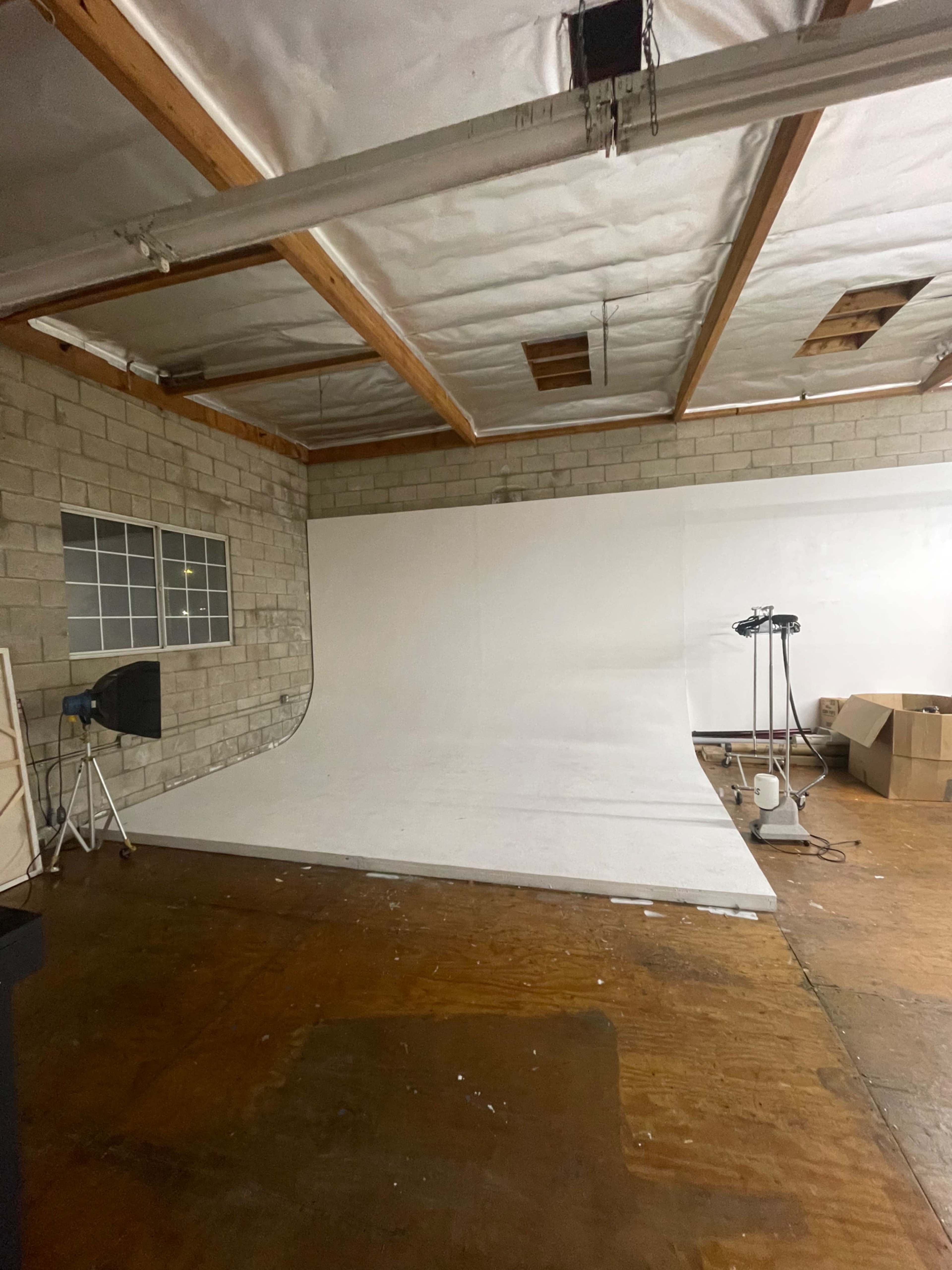 The image shows a spacious room with a white backdrop and a wooden floor, equipped with studio lighting and a few boxes in the corner.
