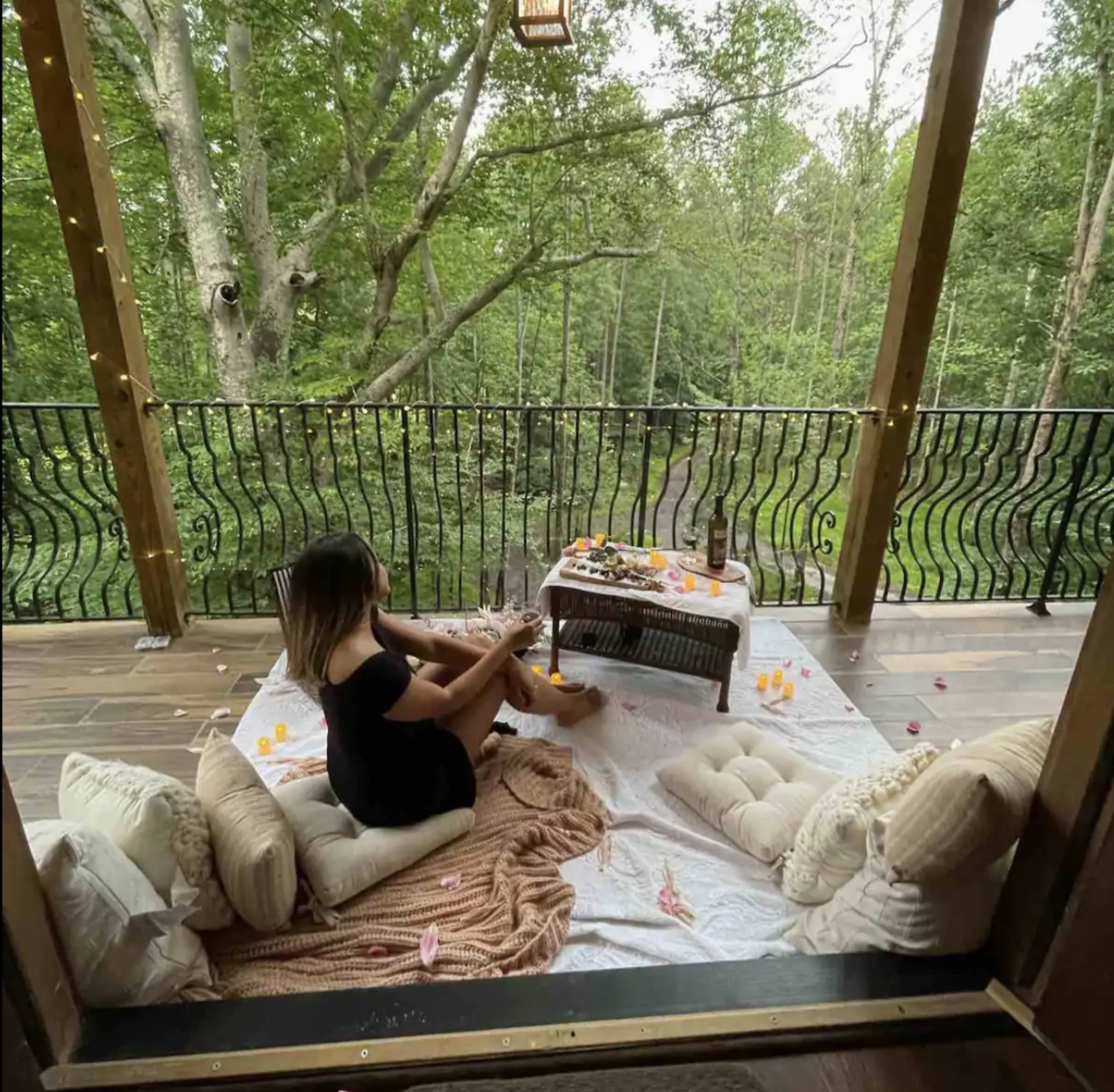 A person sits on a white blanket surrounded by cushions on a wooden deck overlooking a forested area.
