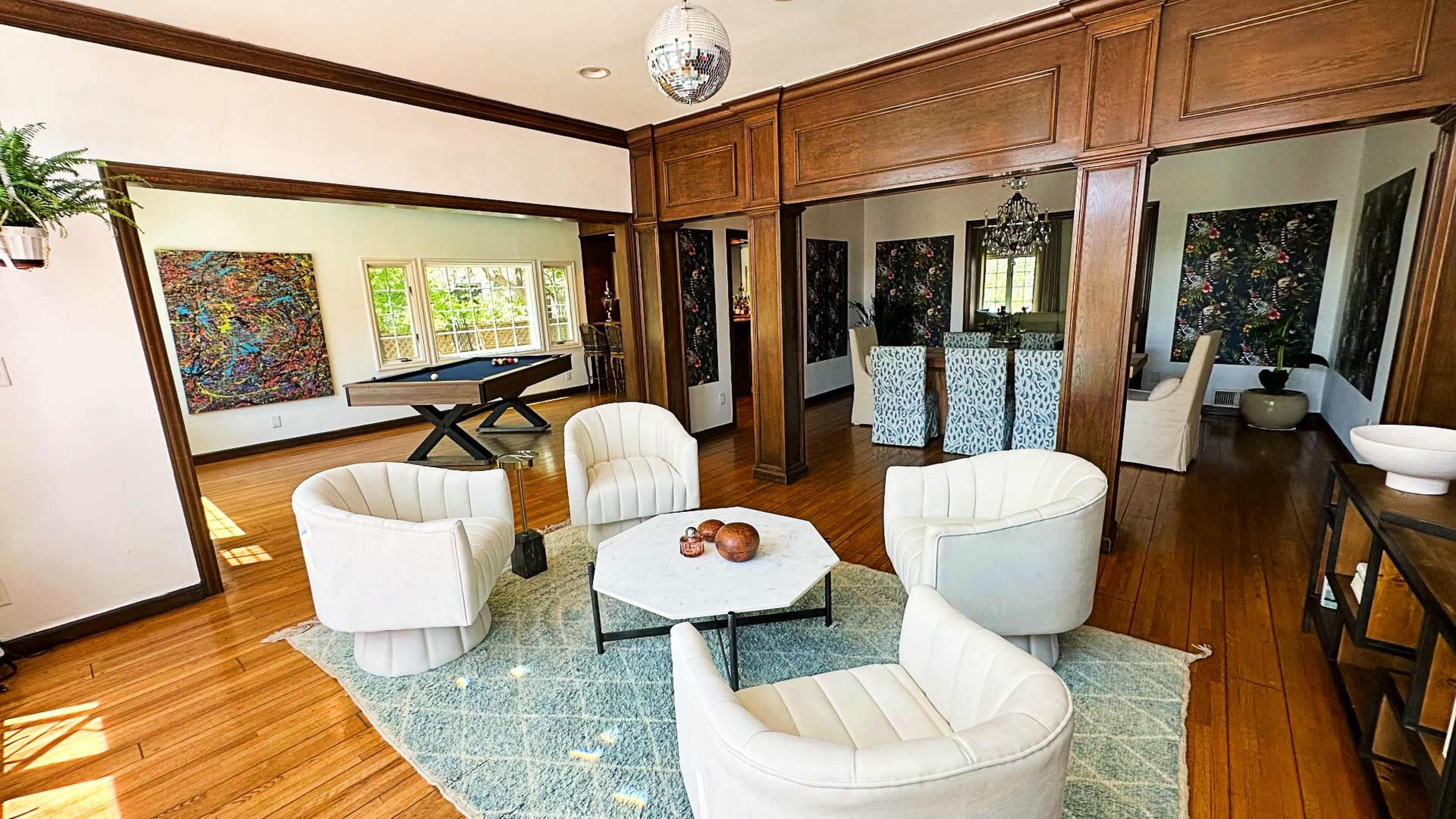 A spacious living room with four white chairs arranged around a coffee table, flanked by two abstract artworks and a pool table in the background.