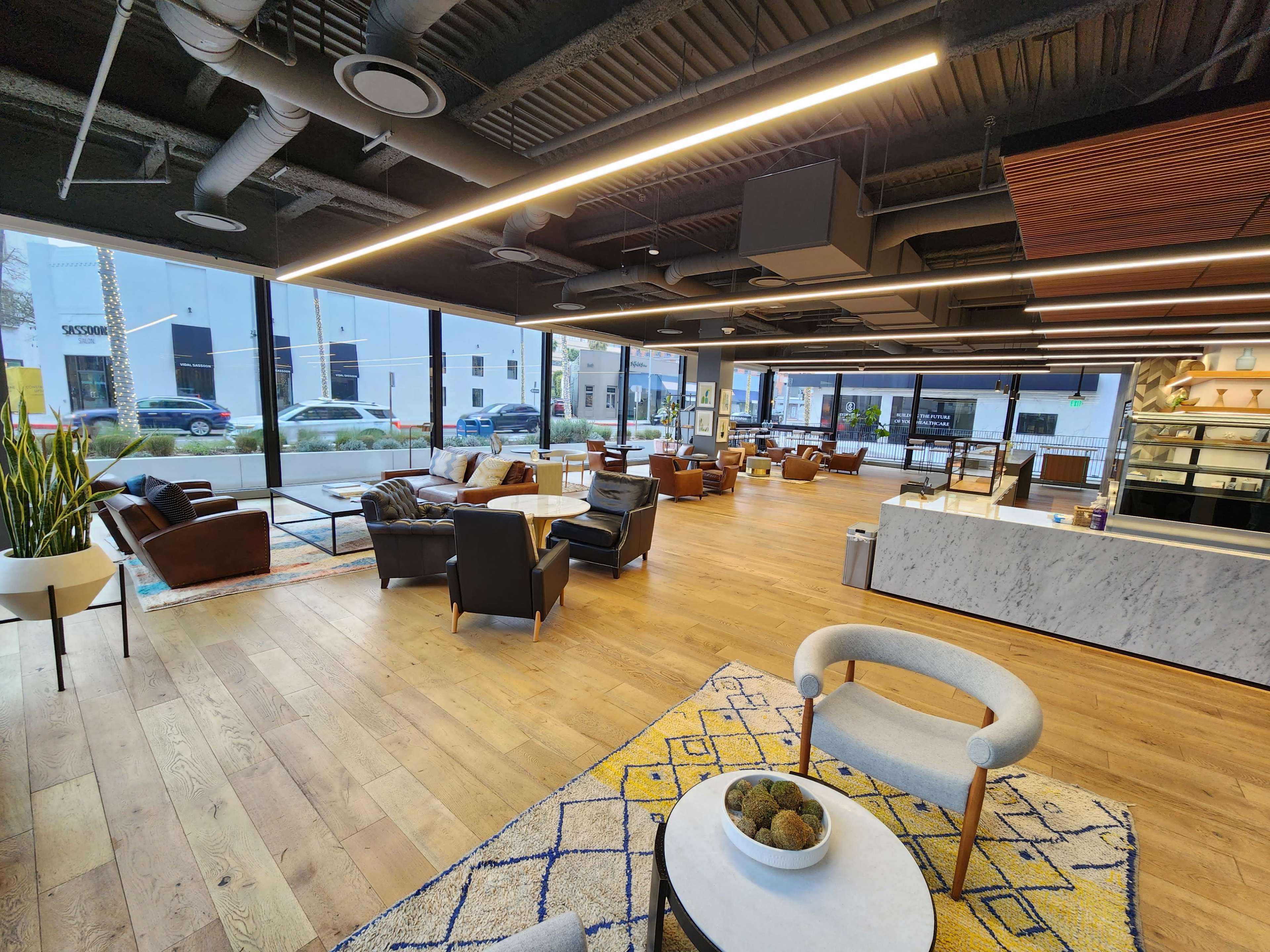 A spacious, modern office lounge with wooden floors, large windows, and a mix of seating arrangements.