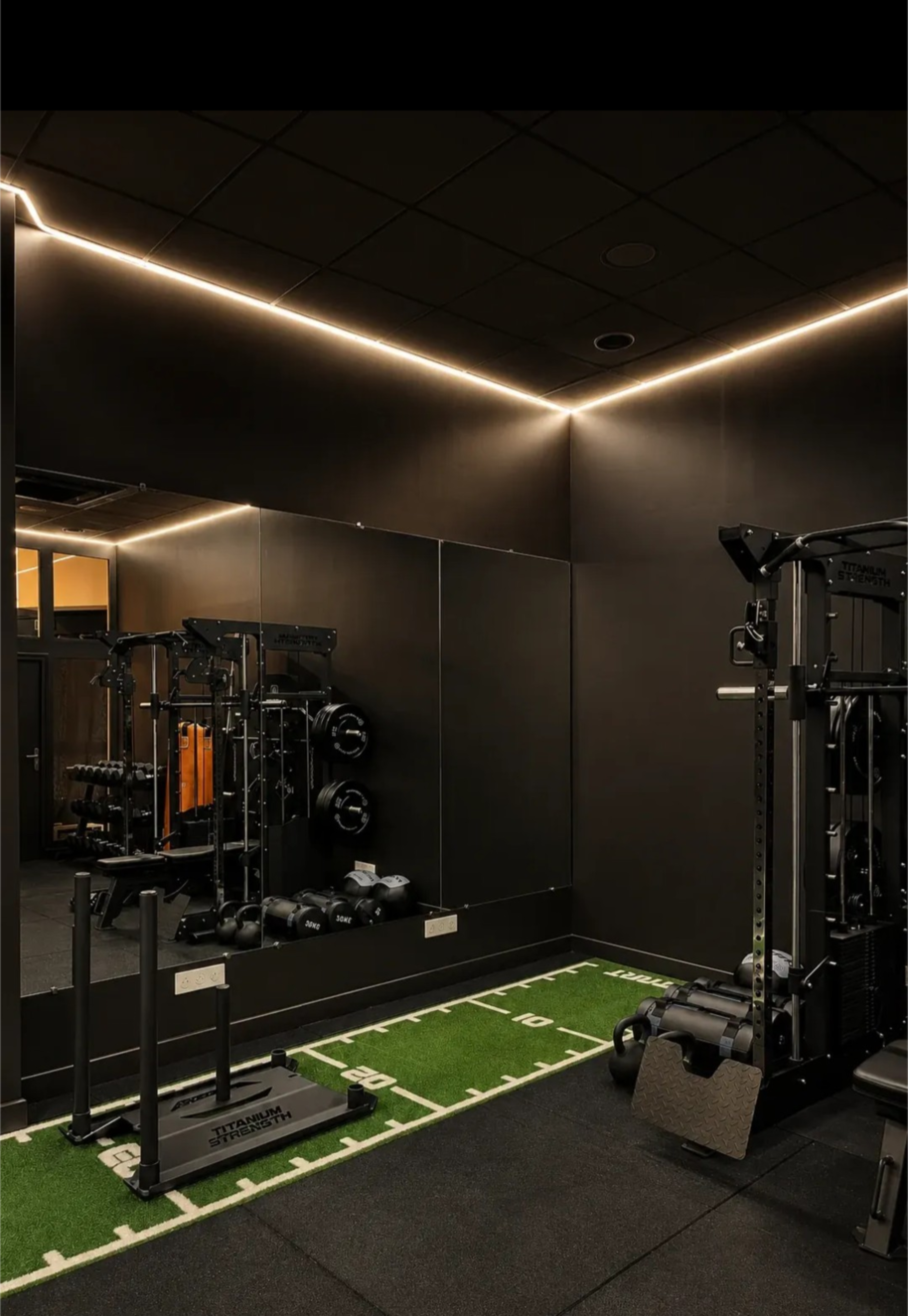 The image shows a modern gym with black walls, a large mirror, and a turf area designed for training, featuring various equipment like free weights and a multi-station strength machine.