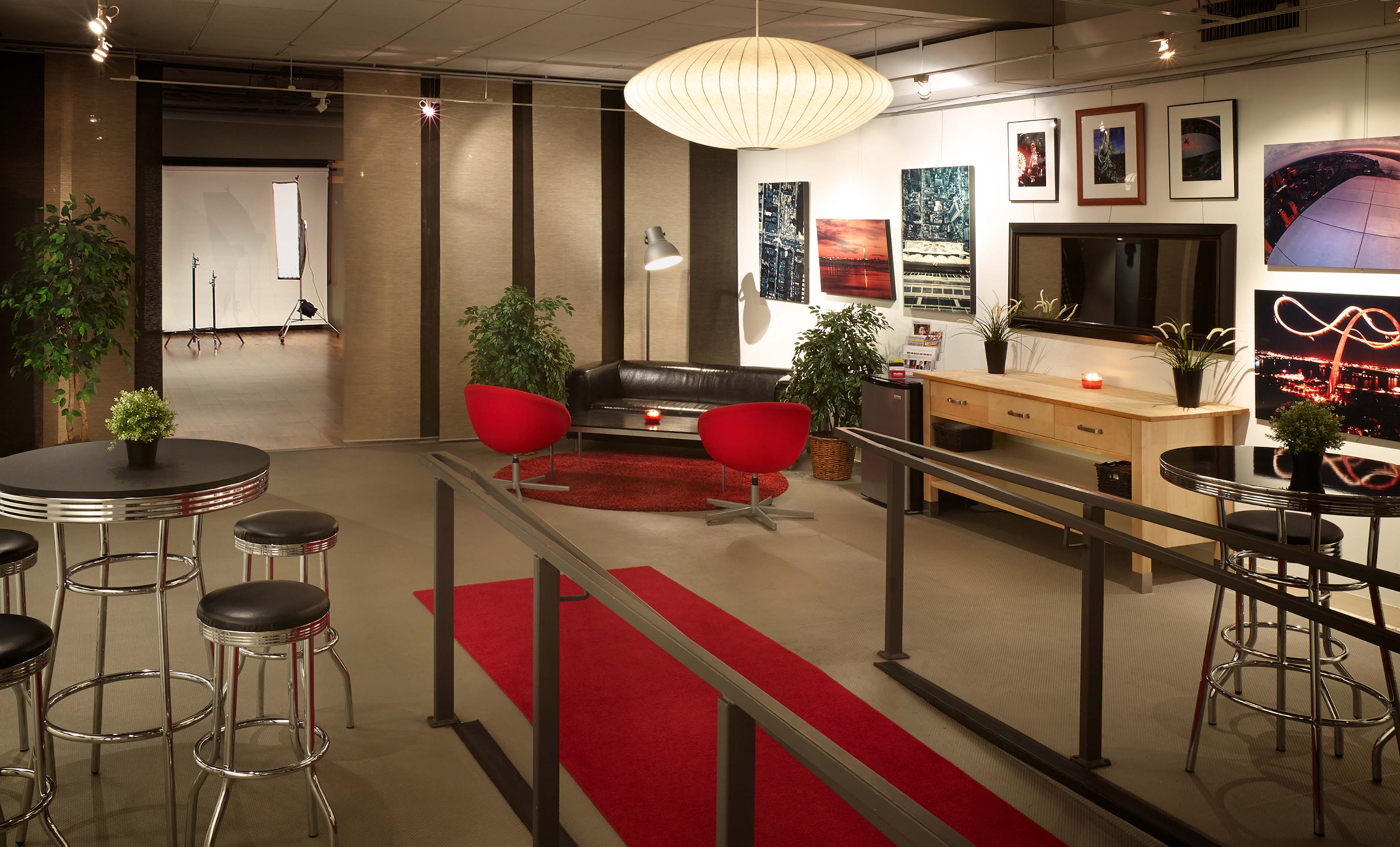 A modern lounge area with a black sofa, red accent chairs, a coffee table, and framed photos on the walls, illuminated by pendant lighting.