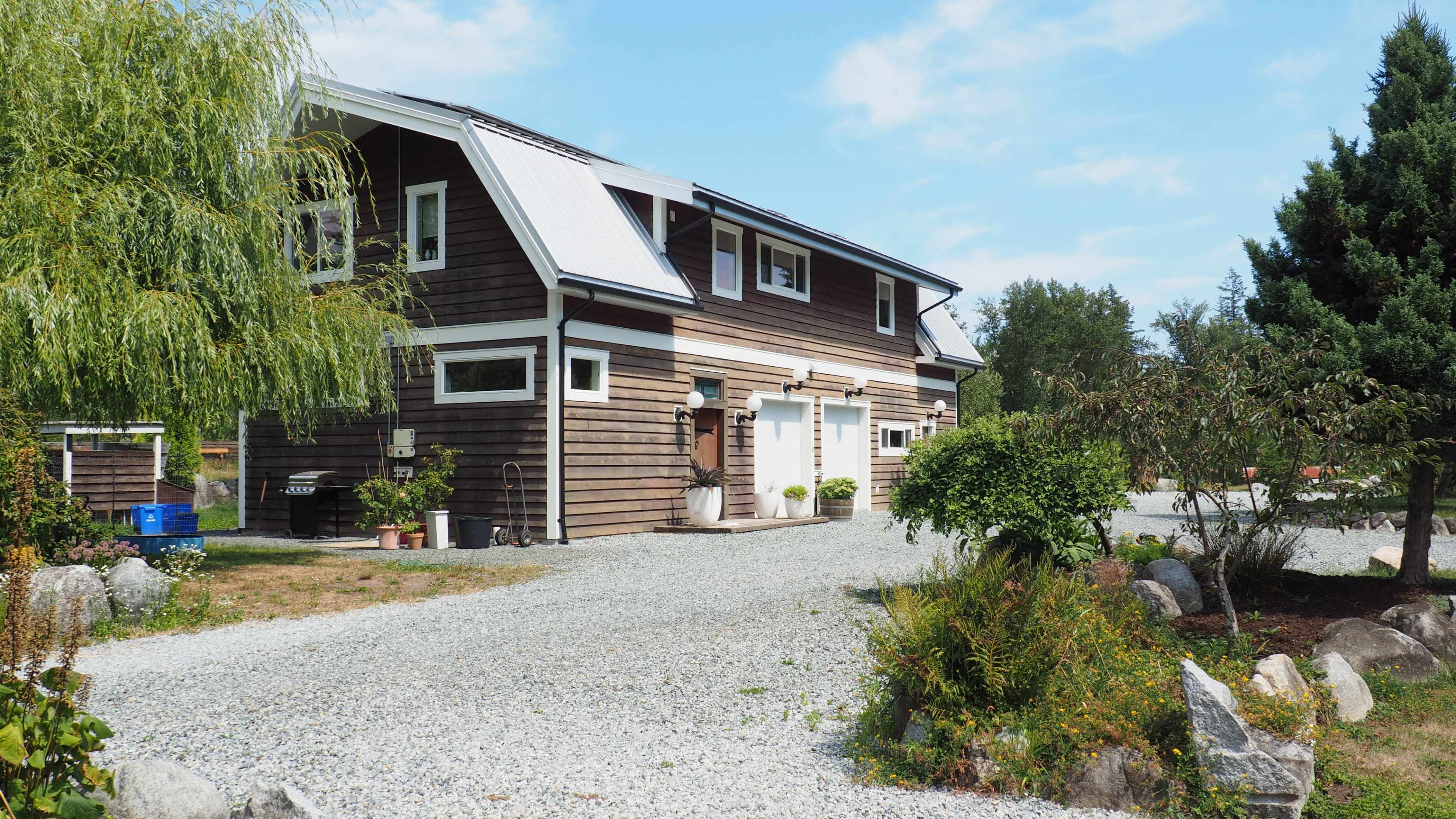 A modern two-story house with a wooden facade is set on a gravel driveway surrounded by landscaped greenery.