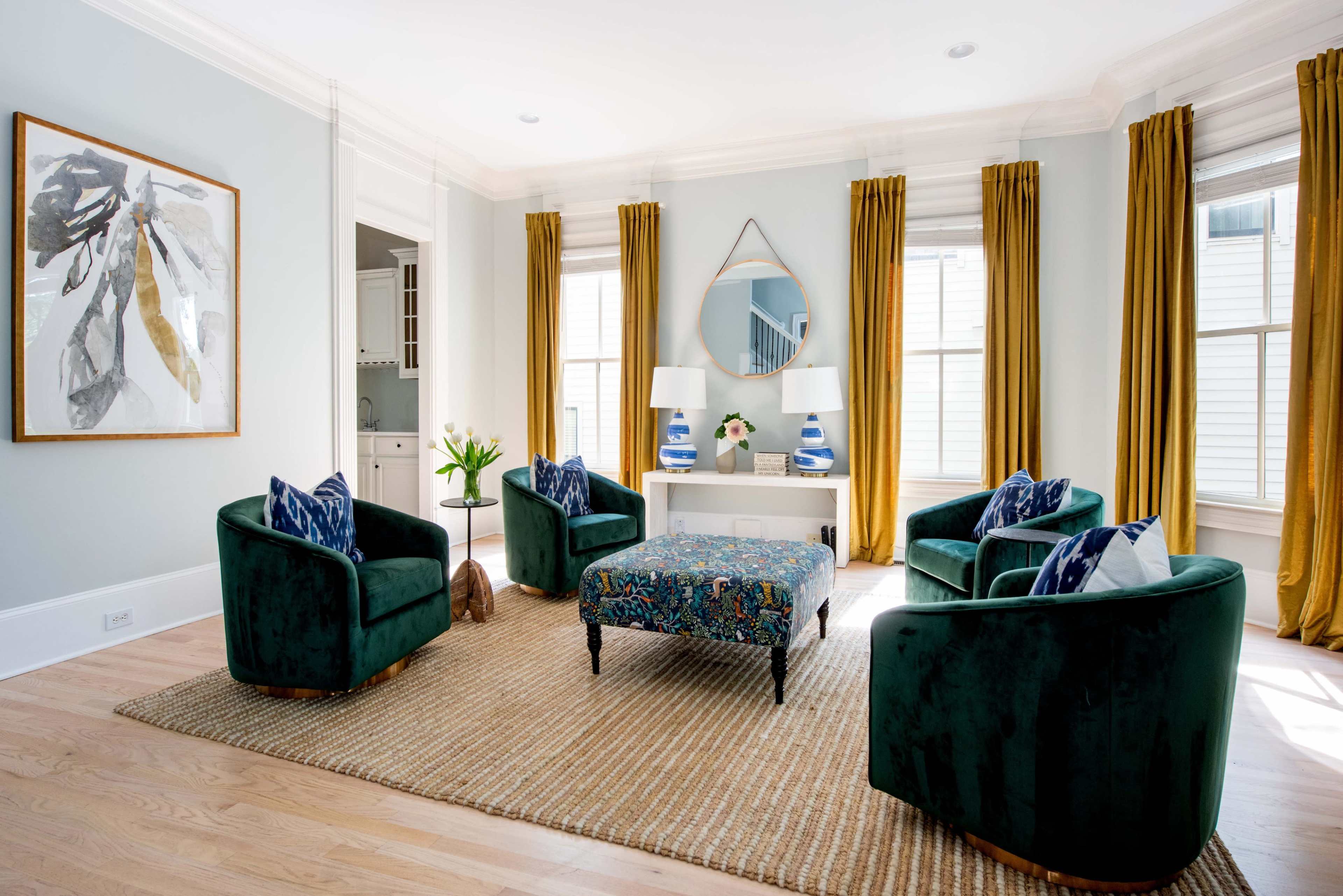 The image shows a bright living room featuring four green armchairs, a patterned ottoman, a large mirror, and two lamps on a console table, surrounded by large windows with golden drapes.