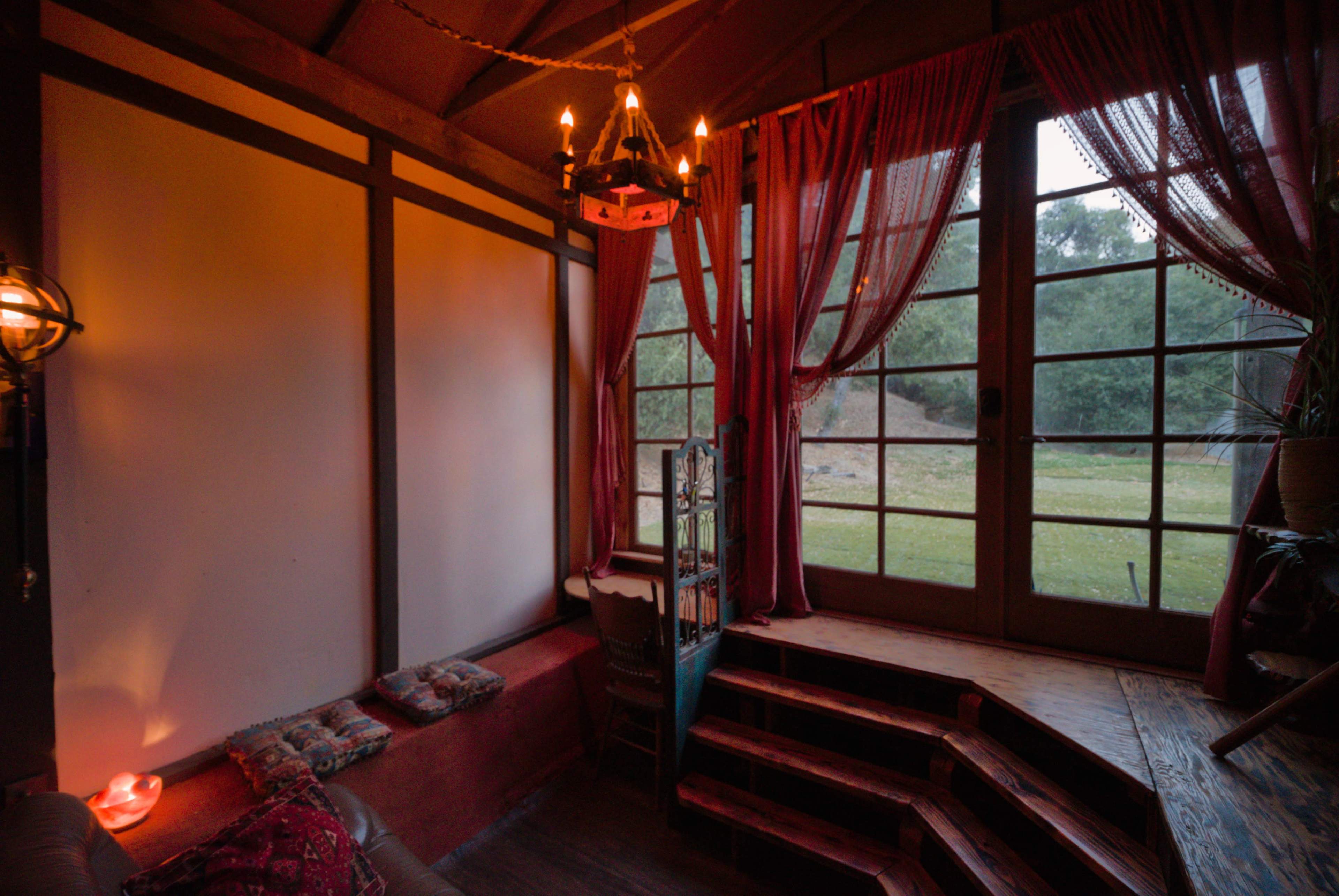 The image shows a cozy interior space with large windows, soft lighting, and wooden steps leading down to a grassy area outside.