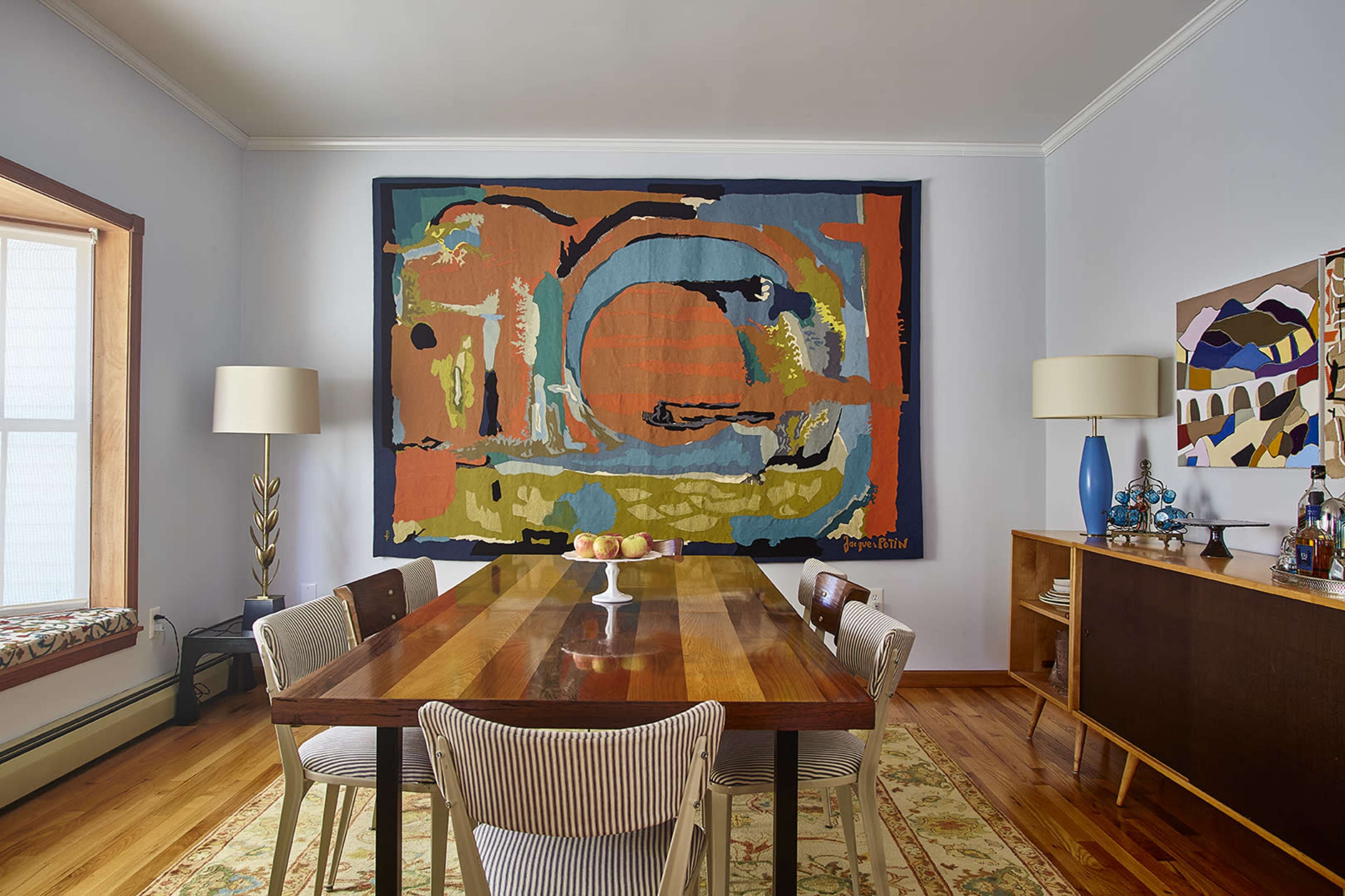 The scene shows a dining room with a wooden table surrounded by striped chairs, a fruit bowl in the center, and colorful wall art above it.