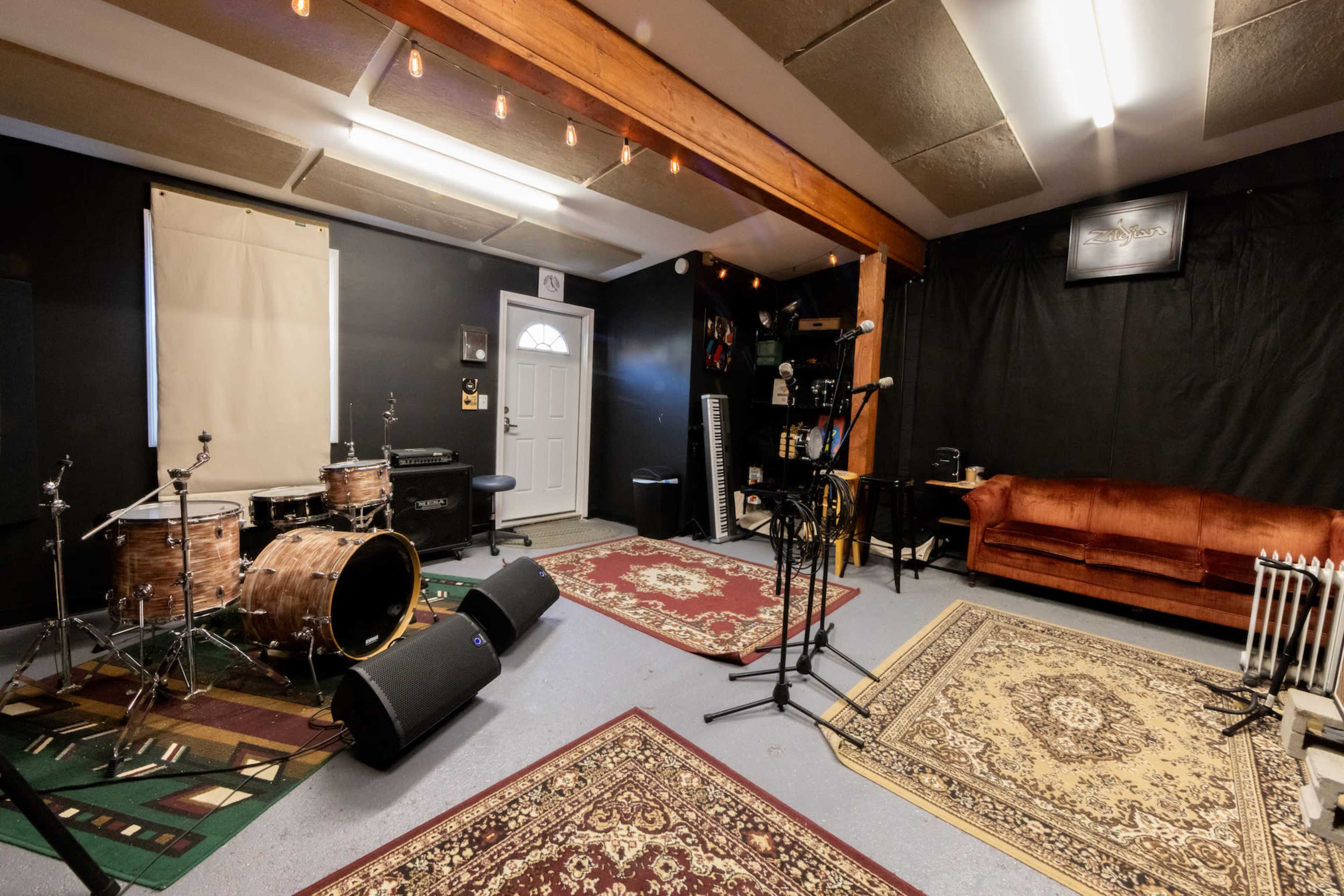 A music rehearsal space features a set of drums, microphones on stands, a keyboard, and a brown couch, all set against dark walls and a door leading outside.