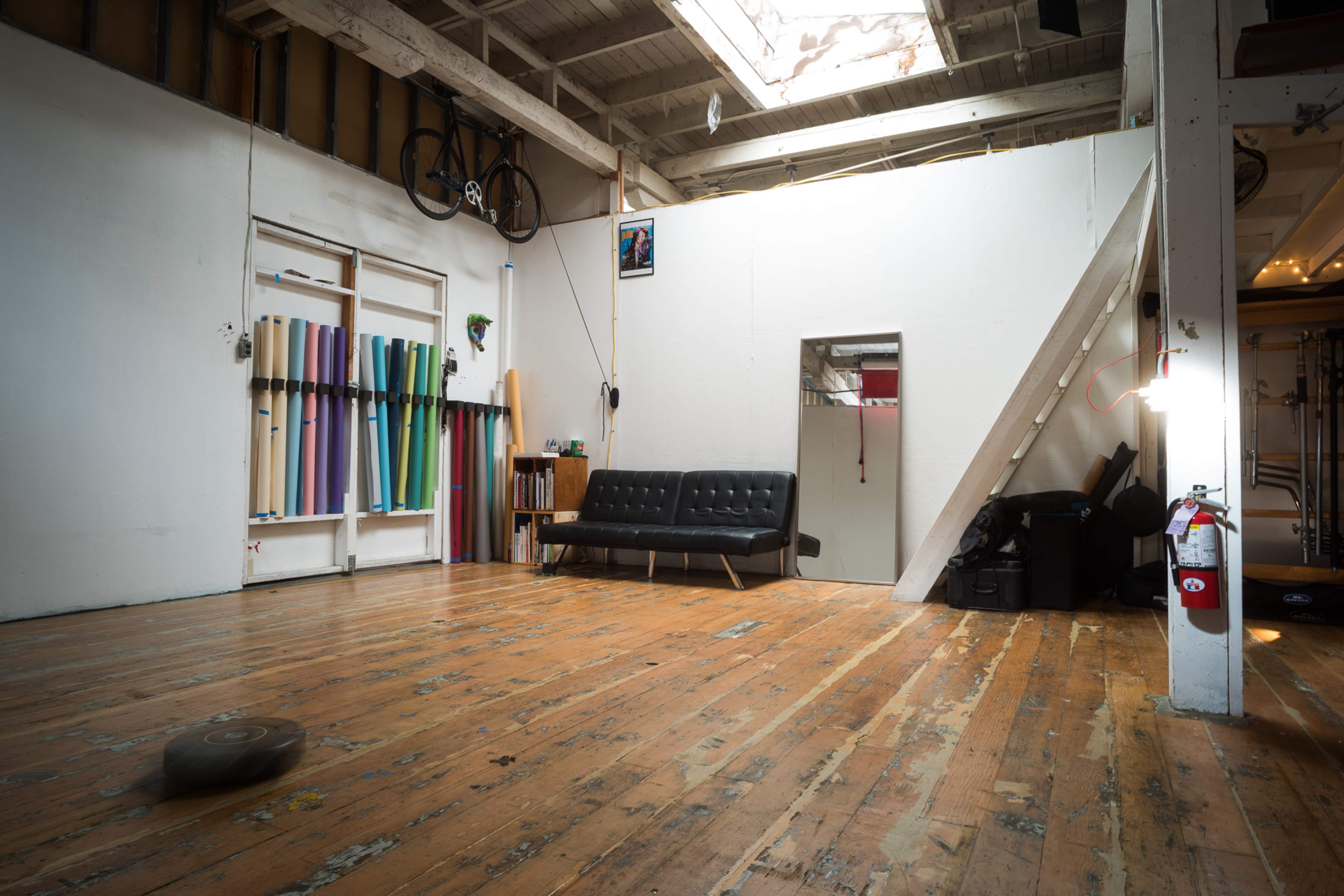 An empty, well-lit room with wooden floors, a black sofa, a bicycle hanging on the wall, and various colored rolls of material displayed along one side.